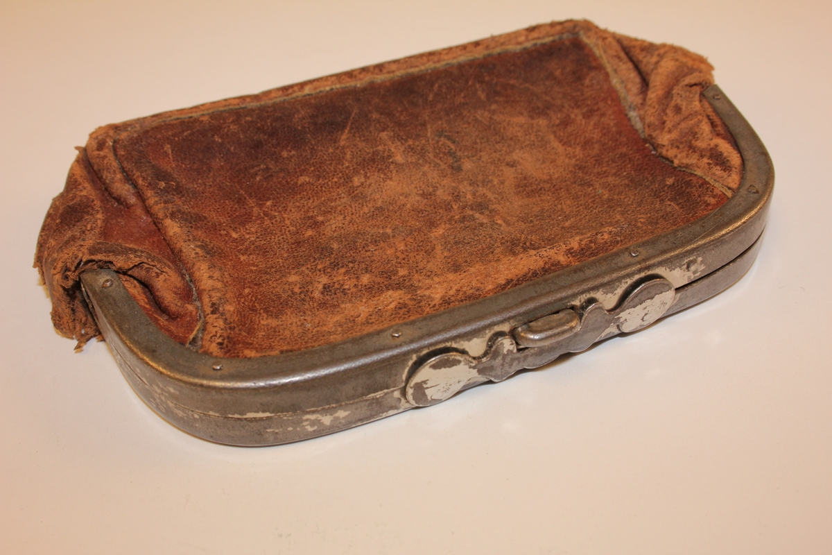 Rektangulær/oval lommebok i skinn. Hengsla metallbøyle med knapplås. Innvendig hengsla metallbøyle som har delt lommeboka i to rom. Skinnet som har fungert som skillevegg, festa til innvendig hengsla bøyle, er borte. Skinnklaff ved opning (for å lette opning av lommebok).