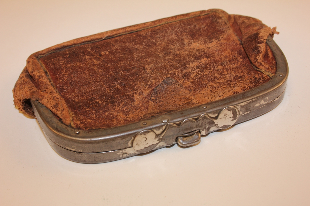 Rektangulær/oval lommebok i skinn. Hengsla metallbøyle med knapplås. Innvendig hengsla metallbøyle som har delt lommeboka i to rom. Skinnet som har fungert som skillevegg, festa til innvendig hengsla bøyle, er borte. Skinnklaff ved opning (for å lette opning av lommebok).