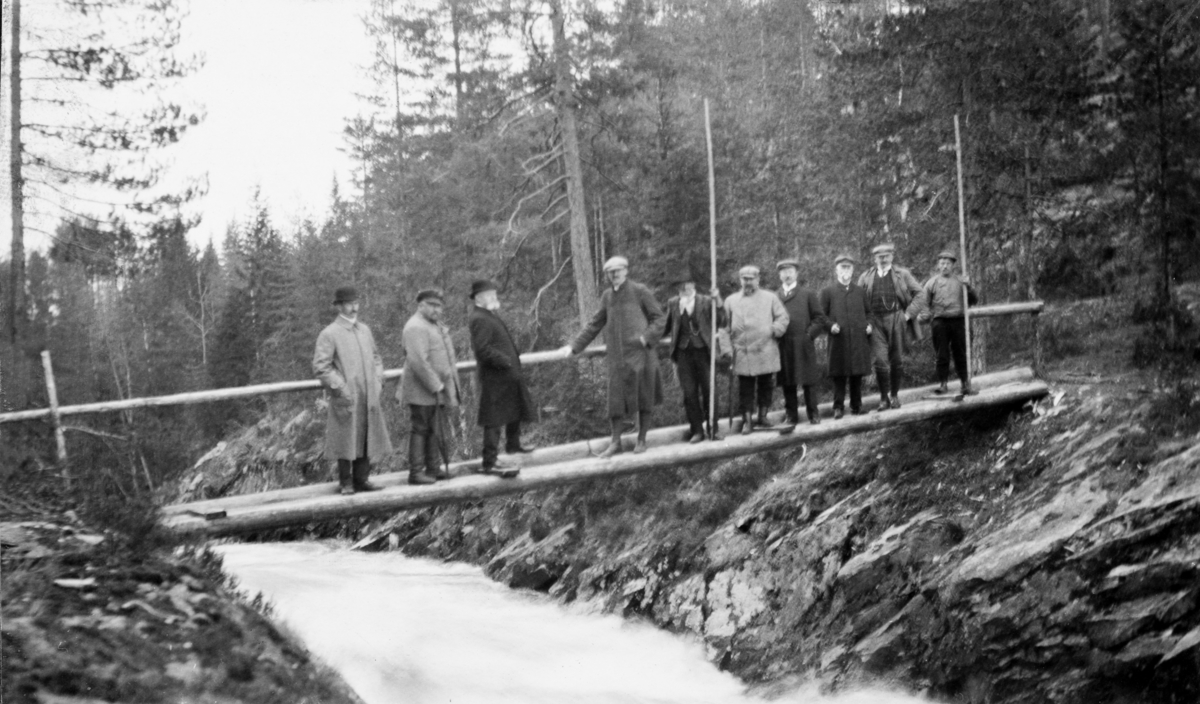 Fløtingsbefaring med Skiensvassdragets Fellesfløtningsforening som vertskap sommeren 1921.  Fotografiet viser ti menn på ei bru over et smalt, men strømrikt bekkeløp i bergrikt lende.  To av karene holder i lange fløterhaker, og var antakelig stedlige tømmerfløtere.  De øvrige dress- og frakkekledde karene var antakelig ledere fra fellesfløtingsforeninger i andre norske vassdrag. 