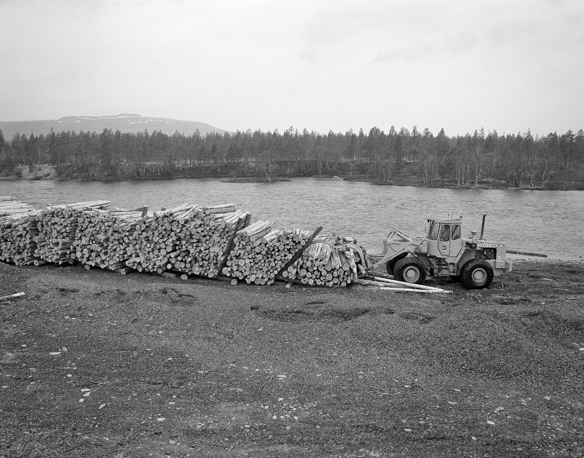 Utislag med hjullaster fra tømmerterminalen ved Sølenstua, øverst i Trysilvassdraget. Fra 1965 og framover fikk Klarälvens Flottningsförening opparbeidet flere terminalplasser av den typen vi ser på dette fotografiet i Trysil og Engerdal.  Slike løsninger ble mulige etter at skogsbilvegnettet i de to kommunene var blitt såpass stort at man anså det for mulig å avvikle fløtinga i de minste sidevassdragene. Omlegginga skjedde i forståelse med Trysilvassdragets skogeierforening.  Terminalplassene hadde ikke bare hjullastere, men i mange tilfeller også barkemaskiner, noe som overflødiggjorde det manuelle arbeidet med barkespader, og dermed forenklet driftsprosessene på hogstfeltene vinterstid.