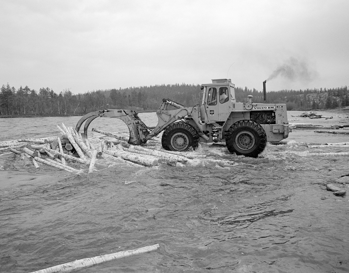 Utislag med hjullaster fra en terminalplass ved Sølenstua i Engerdal i Hedmark. Tømmeret hentes på land, der det var lagt opp ved bredden av Femundselva (Trysilelva).  Med den digre, frontmonterte, hydrauliske kloa som arbeidsredskap hentet maskinføreren tømmerstokkene, som han deretter slapp ut i elva med sikte på at de skulle flyte videre med strømmen. Dette fotografiet ble tatt 22. mai 1986.  Tømmerterminalen på Røskjota ble bygd etter at Klarälvens Flottningsörening i 1953-54 bidro til at det ble bygd bilveger mellom Sølenstua og Galten, og opp til Femundsenden.  Dermed kunne tømmer fra Femund tas opp ved Gløta, og virke fra traktene rundt Isteren kunne hentes ved Isterfossen og kjøres til denne velteplassen, hvor stokkene ble lagt opp for tørking og fløting påfølgende år.  Fra 1965 og framover fikk Klarälvens Flottningsförening opparbeidet flere terminalplasser av den typen vi ser på dette fotografiet i Trysil og Engerdal.  Slike løsninger ble mulige etter at skogsbilvegnettet i de to kommunene var blitt såpass stort at man anså det for mulig å avvikle fløtinga i de minste sidevassdragene. Omlegginga skjedde i forståelse med Trysilvassdragets skogeierforening.  Terminalplassene hadde ikke bare hjullastere, men i mange tilfeller også barkemaskiner, noe som overflødiggjorde det manuelle arbeidet med barkespader, og dermed forenklet driftsprosessene på hogstfeltene vinterstid.

Hjullasteren som ble brukt da dette fotografiet ble tatt var en Volvo BM LM 1240.  Denne modellen ble produsert i perioden 1972-79.  Den veide 12,1 tonn og kunne ta 7-8 tonn i ett grep.  Under utislaget ved Femundselva i 1986 ble det brukt tre hjullastere som alle tilhørte maskinentreprenøren og rallyføreren Olle Arnesson fra Likenäs i Torsby kommune i Sverige.  Maskinførere var Pinnochio Carlson, Eber Halvardsson og Ove Jonsson.