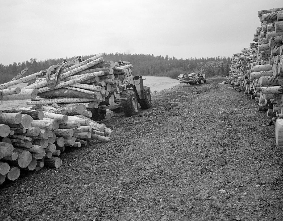 Utislag med to hjullastere fra en terminalplass på Røskjota ved Sølenstua i Engerdal i Hedmark. Tømmeret ble hentet på elvebakken, der det var lagt opp i velter langs bredden av Femundselva (Trysilelva).  Med de digre, frontmonterte, hydrauliske klørne som arbeidsredskaper løftet og kjørte maskinførerne tømmerstokkene, som de deretter slapp ut i elva med sikte på at det skulle flyte videre med strømmen. Dette fotografiet ble tatt 22. mai 1986.  Tømmerterminalen på Røskjota ble bygd etter at Klarälvens Flottningsörening i 1953-54 bidro til at det ble bygd bilveger mellom Sølenstua og Galten, og opp til Femundsenden.  Dermed kunne tømmer fra Femund tas opp ved Gløta, og virke fra traktene rundt Isteren kunne hentes ved Isterfossen og kjøres til denne velteplassen, hvor stokkene ble lagt opp for tørking og fløting påfølgende år.  Fra 1965 og framover fikk Klarälvens Flottningsförening opparbeidet flere terminalplasser av den typen vi ser på dette fotografiet i Trysil og Engerdal.  Slike løsninger ble mulige etter at skogsbilvegnettet i de to kommunene var blitt såpass stort at man anså det for mulig å avvikle fløtinga i de minste sidevassdragene. Omlegginga skjedde i forståelse med Trysilvassdragets skogeierforening.  Terminalplassene hadde ikke bare hjullastere, men i mange tilfeller også barkemaskiner, noe som overflødiggjorde det manuelle arbeidet med barkespader, og dermed forenklet driftsprosessene på hogstfeltene vinterstid.