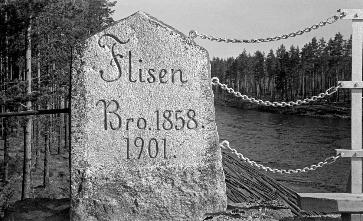 Steinstøtte med teksten «Flisen Bro. 1858. 1901».  Støtta er en flat stein, som er reist på høykant, med et topparti som er formet som et saltak.  Den gjengitte teksten er hogd inn i steinen på den sideflata som vendte mot dem passerte på vegen, antakelig for å markere bruas bygge- og ombyggingsår.  Da dette fotografiet ble tatt hadde man, tilsynelatende nylig, satt inn tre øyeskruer i kanten av støtta.  Fra disse øyeskruene gikk det tre korte kjettinger til hver av de horisontale stagene i om rekkverket på brua.  Fotografiet ble tatt i 1950, og det var antakelig da at støtta fikk den beskrevne forankringa.  I bakgrunnen ser vei ei forholdsvis bred, stilleflytende elv, med en del innlenset tømmer langs den ene elvebredden.  Omkring elva vokste det furuskog.