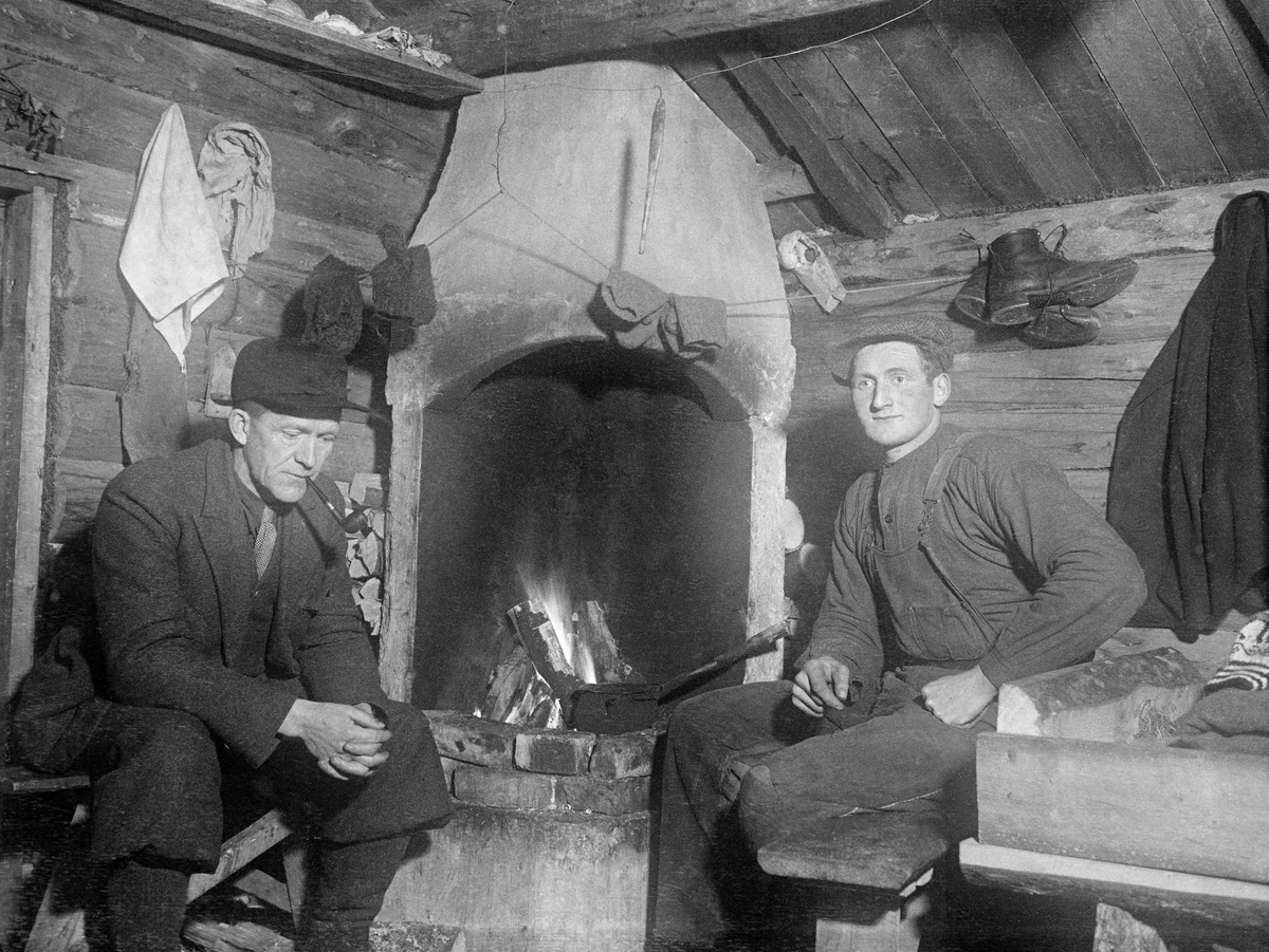 Koieinteriør med peis, antakelig fotografert i andre halvdel av 1920-åra.  Den nevnte peisen var oppført i hjørnet av det laftete rommet, men i såpass avstand fra tømmerveggene at det var rom for et lite vedlager på baksida.    Peiskappa hadde en rundbuet form.  Det var varme på ildstedet, hvor vi også skimter ei gryte.  Framfor peisen satt det to karer.  Til venstre ser vi en ung mann, kledd i overalls av bomull med et busserulliknende plagg på overkroppen og sixpencelue på hodet.  Dette var antakelig en skogsarbeider.  Han satt på en krakk ved enden av ei seng.  På veggen bak ham hang det ei  mørk jakke og et støvlepar.  Til venstre i bildet satt en middelaldrende mann på  en krakk ved døra.  Han var kledd i nikkersdress av vadmel, hadde slips i halsen og ei mørk østerdalslue på hodet og ei tobakkspipe i munnen.  Ut fra påkledningen er det nærliggende å anta at dette var skogeieren eller en skogfunksjonær.

Fotografiet er tatt i forbindelse med undersøkelser forstkandidat Johan Sønnik Andersen gjorde i Hedmark, Oppland og Nord-Trøndelag i andre halvdel av 1920-åra.  Observasjonene og refleksjonene hans ble utgitt under tittelen «Undersøkelse av skogshytter og skogsarbeidernes levevilkår» i 1932.  

Norsk Skogmuseum har overtatt en del av de fotografiske negativene fra Sønnik Andersens undersøkelser i Hedmark, Oppland og Nord-Trøndelag.  Noen få av disse er brukt som illustrasjoner i det nevnte heftet.  De er imidlertid – antakelig for å sikre involverte skogeiere og skogsarbeidere en viss anonymitet – svært knapt tekstet fra opphavsmannens side.  Dette har naturligvis også fått konsekvenser for hvordan Norsk Skogmuseum kan registrere motivene i sin fotodatabase.