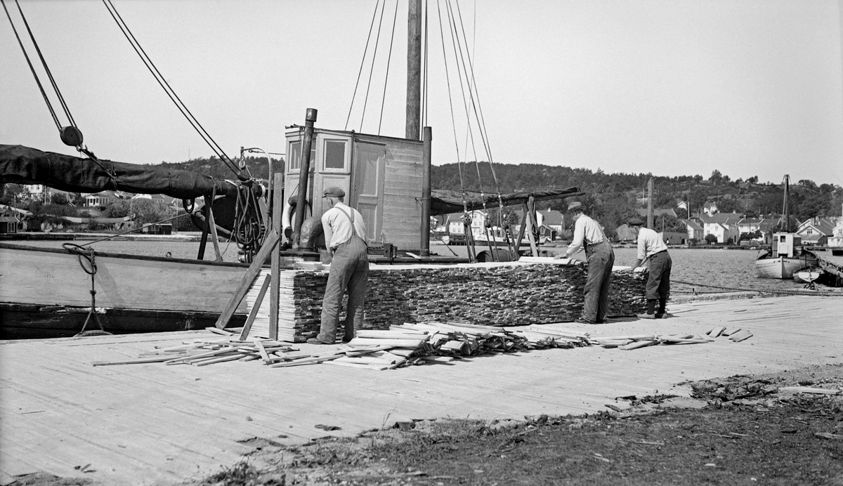 Lasting av stavved på havna i Lillesand i Aust-Agder sommeren 1935.  Veden lå stablet ved kaikanten, der tre menn plukket dem opp for å bære dem over i ei bakenforliggende skøyte med motor og rigg for seiling.  Det er i dette tilfellet noe uklart om staven skulle brukes til parkett- eller tønneproduksjon.