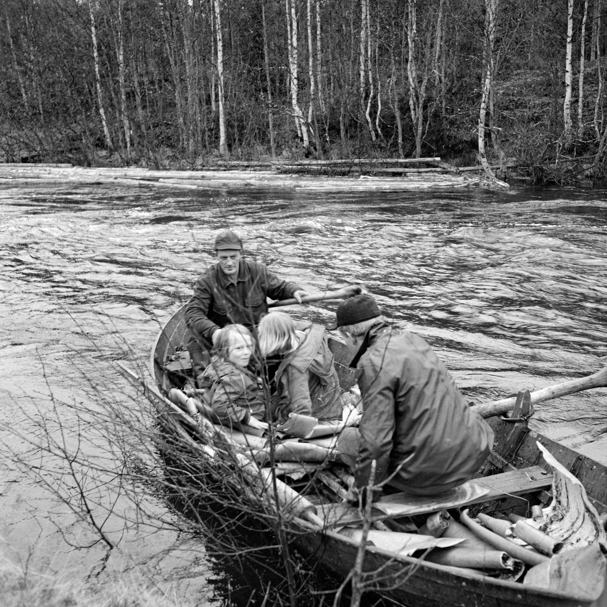 Fra tømmerfløtinga i elva Julussa, som renner gjennom deler av Elverum og Åmot kommuner i Hedmark. Bildet oppgis å være tatt i 1967. Det viser en fløterbåt med to mann og to små jenter om bord. Roeren, som satt med ryggen mot fotografen, er muligens Knut Vold, mens arbeidskameraten som satt i akterenden med ei styreåre minner om Odd Skjærbekk. Jentene i midten av båten er Rønnaug Fossum (datter av museumsmannen som tok bildet) og hennes venninne Inger Langholen. Da fotografiet ble tatt lå det en del neverflak i båten, noe som neppe var vanlig under tømmerfløting. Elva var flomstor, og det lå en del tømmer langs elvebredden i bakgrunnen.