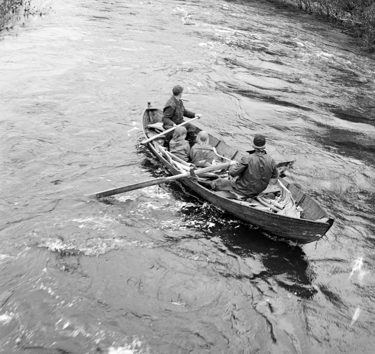 Fra tømmerfløtinga i elva Julussa, som renner gjennom deler av Elverum og Åmot kommuner i Hedmark. Bildet oppgis å være tatt i 1967.  Det viser en fløterbåt med to mann og to små jenter om bord. Roeren, som satt med ryggen mot fotografen, er muligens Knut Vold, mens arbeidskameraten som satt i akterenden med en fløterhake han brukte som styreåre minner om Odd Skjærbekk. Jentene i midten av båten er Rønnaug Fossum (datter av museumsmannen som tok bildet) og hennes venninne Inger Langholen. Da fotografiet ble tatt lå det en del neverflak i båten, noe som neppe var vanlig under tømmerfløting. Elva var flomstor, og det lå en del tømmer langs elvebredden i bakgrunnen.