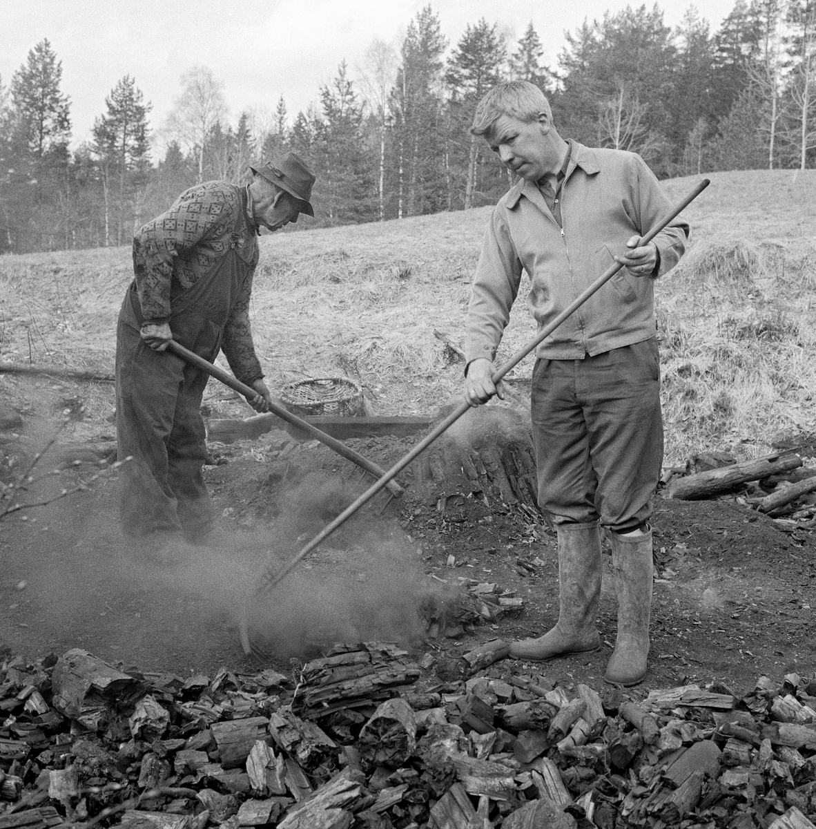 Trekolbrenning (trekullbrenning) hos smeden Jørgen eller Jørn Romundstad (1890-1972) på Vegårshei i Aust-Agder.  Bildet skal være tatt våren 1968.  Vi ser smeden (til venstre) sammen med forstkandidaten og journalisten Andreas Vevstad (1930-2007), som var med museumsbestyrer Tore Fossum som kjentmann da han besøkte Romundstad.  Romundstad og Vevstad rakte kol på milebotnen med langtinnete river.  Begge karene var arbeidskledde.