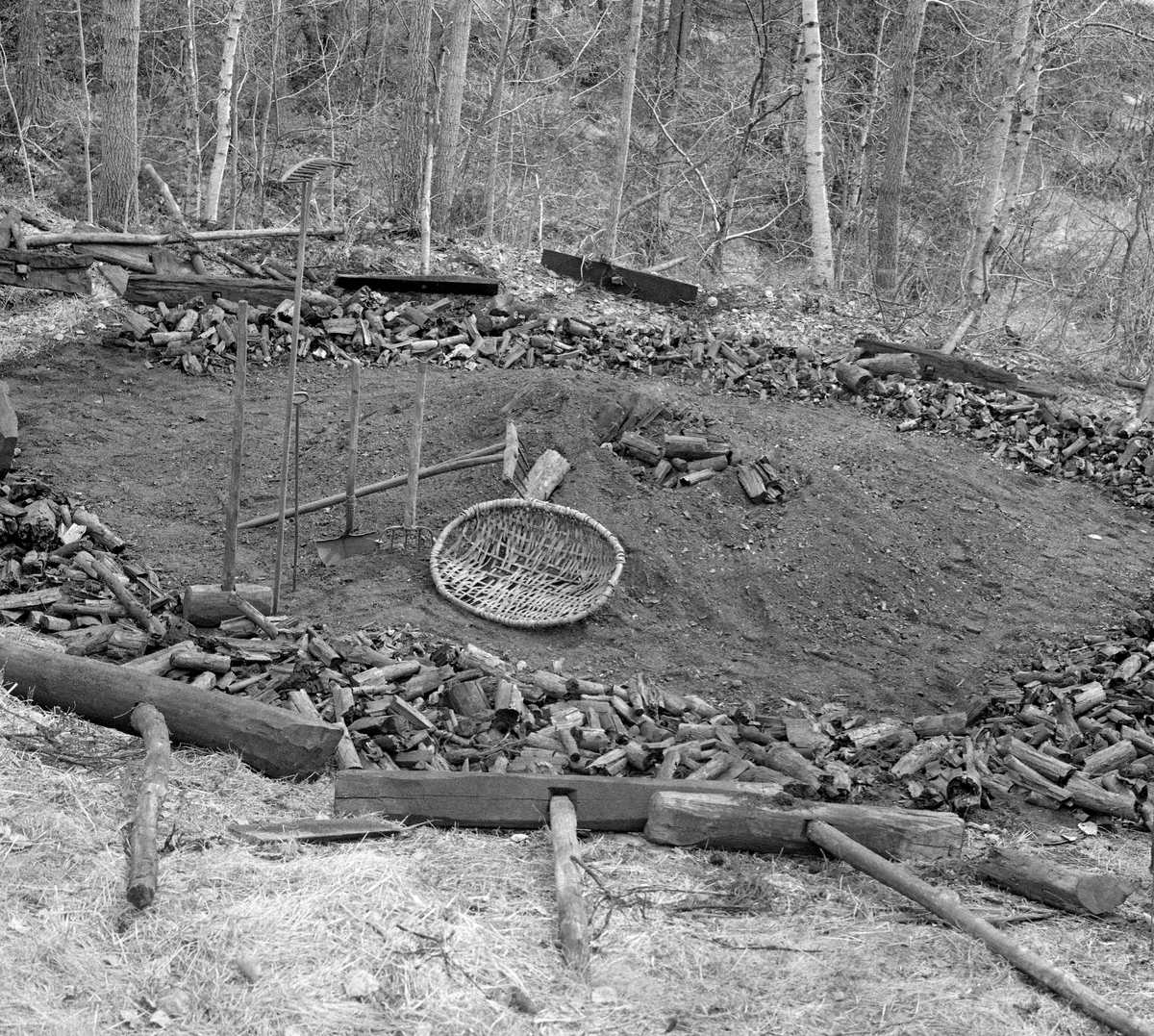 Kolbotn hos smeden Jørgen eller Jørn Romundstad (1890-1972) på Vegårshei i Aust-Agder.  Bildet skal være tatt våren 1968 mot den sirkelformete kolbotnen, der Romundstad hadde rakt kolbitene ut mot sidene.  På opptakstidspunktet lå det bare noen få kolbiter igjen i sentrum.  Framfor lå redskapene som ble brukt under kolbrenninga.  Vi ser et flettet kolfat, et greip, en spade, et spett, to river med litt ulik bredde og tinnelengde og ei treklubbe.  I ytterkanten av kolbotnen lå det stubbehyller, T-formete treelementer som skulle hindre at dekket på de bratte sidekantene av mila raste ned. Ei kort orientering om kolbrenning i slike miler finnes under fanen «Opplysninger».  Kullmile. Mile. Kolmile.