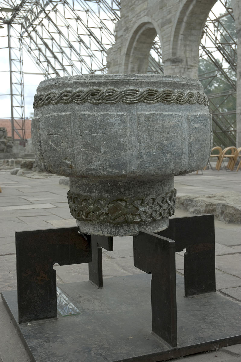 Døpefont fra middelalderen, i kleberstein med flettebord øverst, står på metallstand i Hamardomen, med domkirkeruinen i bakgrunnen.
