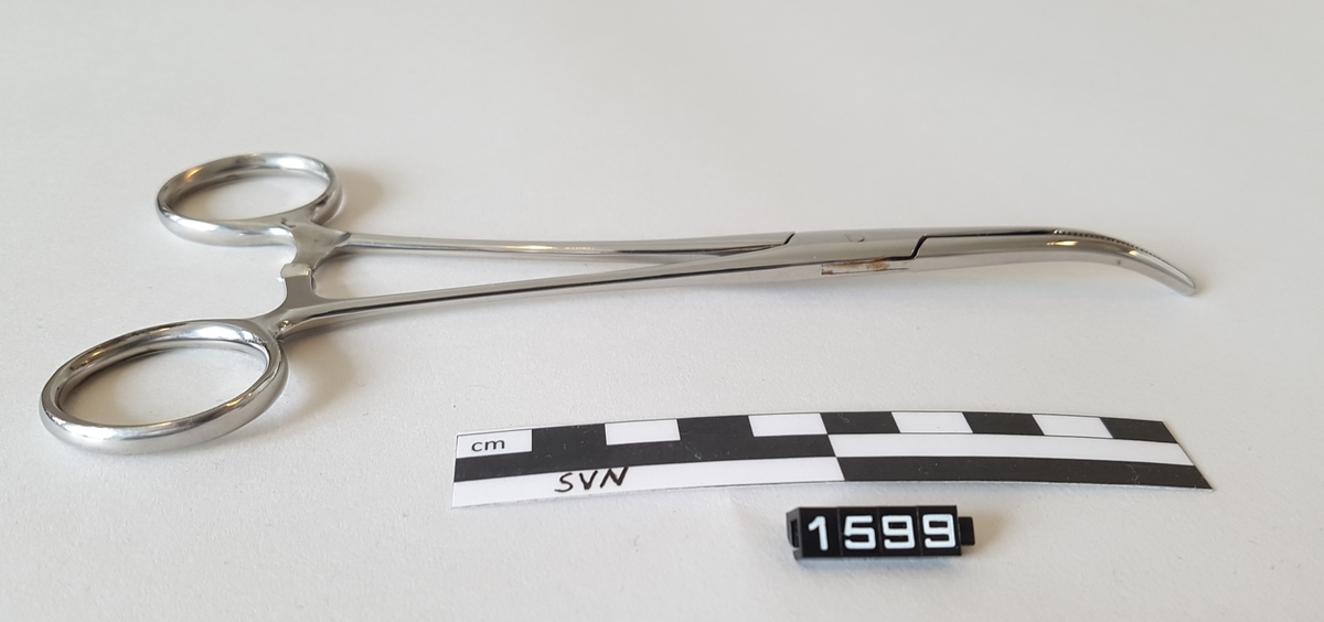 Gjenstanden er en kirurgisk bøyd klemme. 
Klemmen er utformet som en saks, men i stede for skjærekniver er det to flater som møtes og klemmes sammen.
Flatene som møtes har serrateringer, riller. ytterst på klemmen har flatene tenner som går i hverandre.
To tenner på en side og en tann på den andre siden.
Gjenstanden har fingerhull for manipulering av objektet.
like over fingerhullene er det en låsemekanisme med en hake på den ene delen og flere motstående haker på den andre delen
Instrumentet kan låses ved hjelp av disse og hvor mye press det legges på tangspissen bestemmes av hvilken av mothakene 
det låses mot.