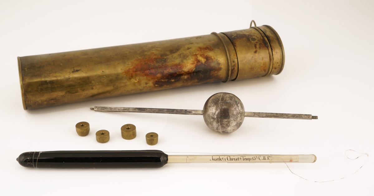 To Alkoholmetere : Ett av antatt sølv, ett av glass  som ligger i et etui med lokk. Lokket inneholder et ekstra rom for oppbevaring av 4 vekter til metallalkoholmeteret. Etuiet inneholder to rom for plassering av alkoholmeterne. Alkoholmeteret av glass er tilført en opphengsanordning av fiskesene.