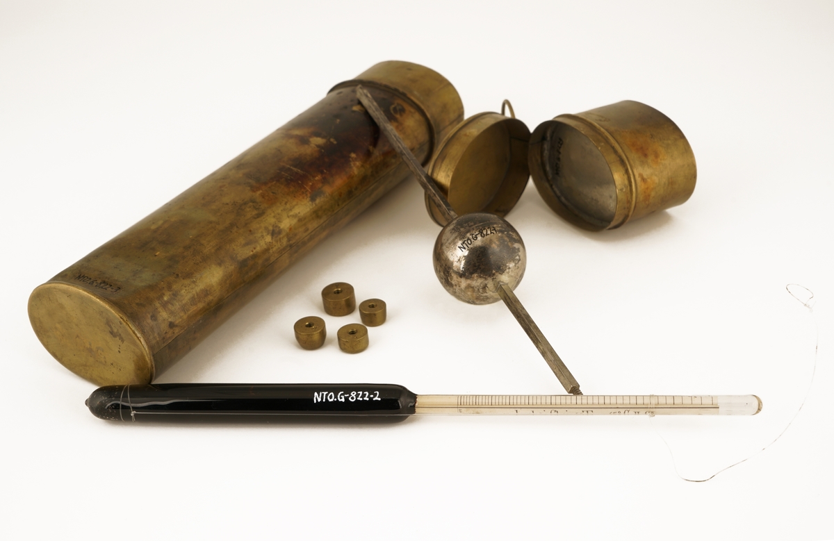 To Alkoholmetere : Ett av antatt sølv, ett av glass  som ligger i et etui med lokk. Lokket inneholder et ekstra rom for oppbevaring av 4 vekter til metallalkoholmeteret. Etuiet inneholder to rom for plassering av alkoholmeterne. Alkoholmeteret av glass er tilført en opphengsanordning av fiskesene.