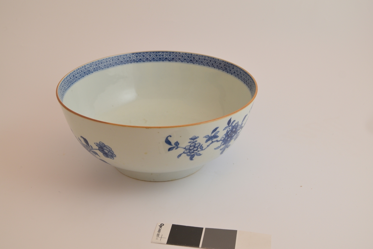 En hvit porselensbolle som er dekorert med blomster på utsiden av bollen og en blå mønstret kant langs toppen på innsiden av bollen som er ca. 2 cm høy. Det er i tillegg in gul stripe langs toppen av veggen til bollen.