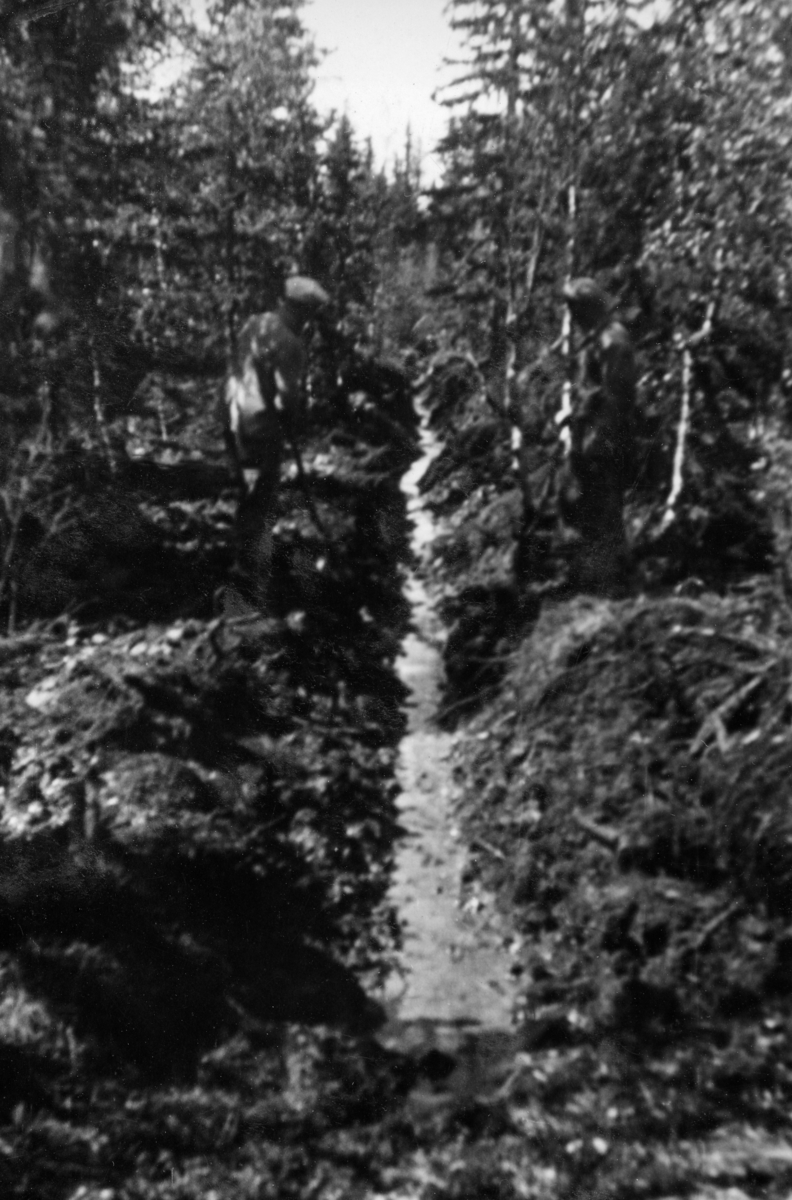 Fra grøftesprengingsprosjekt på skogeiendommen Mustad i Vardal i mai-juni 1948.  Dette fotografiet er det tredje i en serie på tre bilder som viser ulike faser i arbeidet med grøfta.  Vi ser et myrlendt areal hvor det er ryddet en rettlinjet trasé i bjørke- og granskogen.  Her er det foretatt en lineær sprenging av jordmassene i traséens midtlinje, og deretter er løsnet jord, røtter og stein spadd til side slik at vann fra et ovenforliggende sumpareal kunne sive inn i grøfta.  Grøftesprenging var en amerikansk metode som hadde vært prøvd i Norge i 1920-åra uten praktisk suksess.  Før krigen hadde nemlig sprengstoffet vært for kostbart til at denne metoden kunne konkurrere med manuelt arbeid.  Etter krigen ble det lansert en ny type grøftedynamitt i form av patroner som skulle stikkes 20-25 centimeter ned i myra med en innbyrdes avstand på 30-40 centimeter langsetter grøftetraseens midtlinje.  En av patronene ble antent ved hjelp av ei meterlang lunte.  Deretter løp mannskapene unna.  Omtrent et minutt seinere smalt det.  Detoneringa av den første patronen utløste de øvrige, og en vegg av torv, busker og røtter stod til værs - og falt deretter ned på bakken igjen.  Noe falt naturligvis ned i grøftetraseen, men det tok likevel ikke mannskapene mange minuttene å renske grøfta med greip.  Dynamittpatronene var kostbare, de representerte 70-80 prosent av omkostningene, så prisen per meter ferdig grøft ble omtrent den samme ved sprenging som ved manuell grøftegraving.  Med traktorgravernes inntreden utover i 1950- og 60-åra ble metoden stadig mindre aktuell.