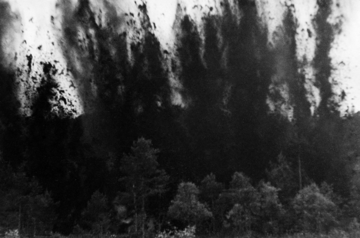 Grøftesprenging, muligens i Løiten Almenning, antakelig sommeren 1949.  Fotografiet viser hvordan jord, røtter og stein ble kastet til værs – høyt over tretoppene i tilstøtende skog – idet salven ble avfyrt.  Grøftesprenging var en amerikansk metode som hadde vært prøvd i Norge i 1920-åra uten praktisk suksess.  Før krigen hadde nemlig sprengstoffet vært for kostbart til at denne metoden kunne konkurrere med manuelt arbeid.  Etter krigen ble det lansert en ny type grøftedynamitt i form av patroner som skulle stikkes 20-25 centimeter ned i myra med en innbyrdes avstand på 30-40 centimeter langsetter grøftetraseens midtlinje.  En av patronene ble antent ved hjelp av ei meterlang lunte.  Deretter løp mannskapene unna.  Omtrent et minutt seinere smalt det.  Detoneringa av den første patronen utløste de øvrige, og en vegg av torv, busker og røtter stod til værs - og falt deretter ned på bakken igjen.  Noe falt naturligvis ned i grøftetraseen, men det tok likevel ikke mannskapene mange minuttene å renske grøfta med greip.  Dynamittpatronene var kostbare, de representerte 70-80 prosent av omkostningene, så prisen per meter ferdig grøft ble omtrent den samme ved sprenging som ved manuell grøftegraving.  Med traktorgravernes inntreden utover i 1950- og 60-åra ble metoden stadig mindre aktuell.  Det fortelles imidlertid at sagbruksarbeiderne i mange år etter at disse forsøkene ble gjort opplevde å få tømmer der steinsplinter fra slike sprengingsforsøk hadde blitt skutt inn i veden på sagbenkene.  Dette var ikke helt ufarlig.