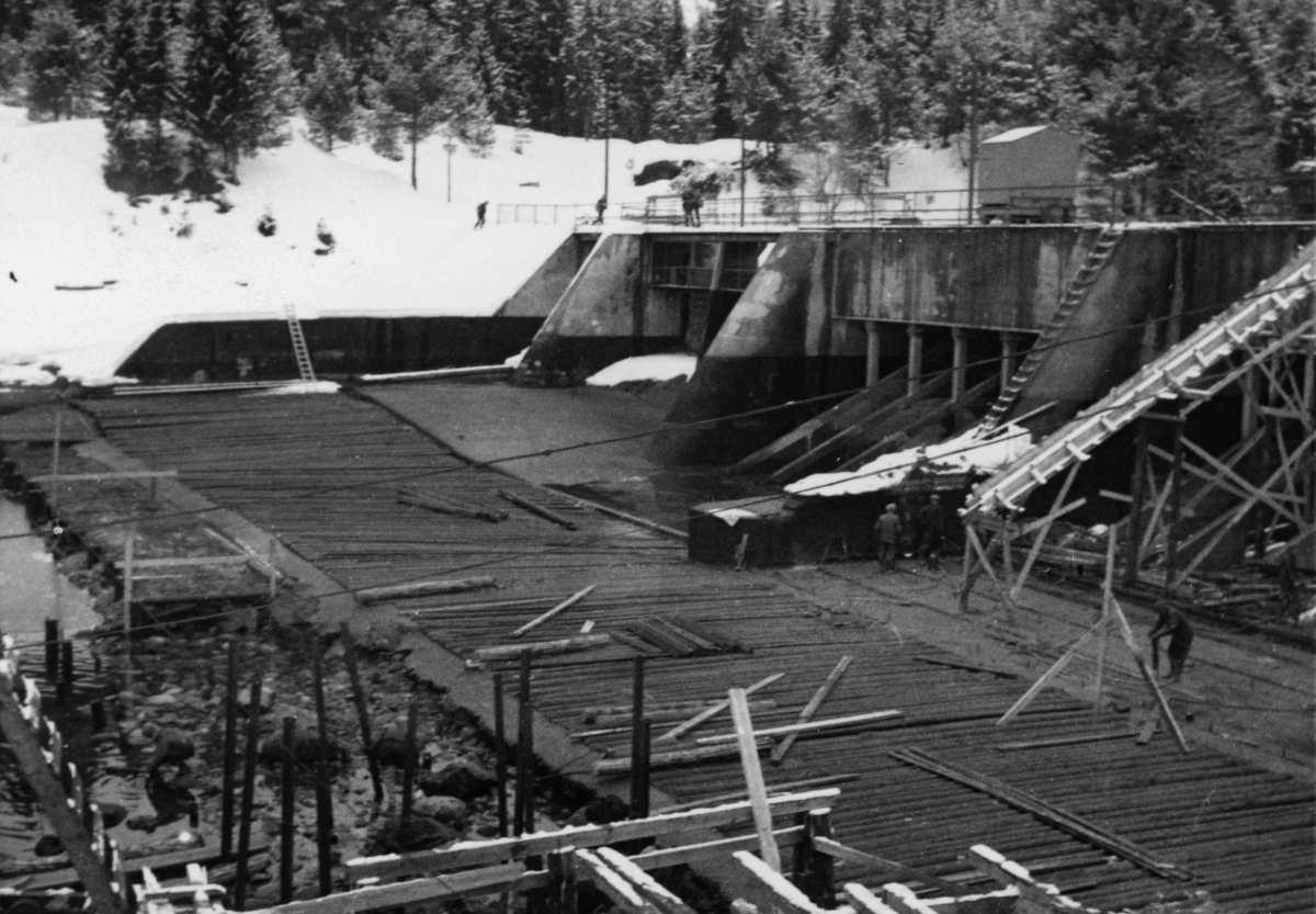 Damarbeid ved Tinnoset på seinvinteren, antakelig omkring 1960.  Bildet er tatt mot den golvlagte elvebotnen like nedenfor damkonstruksjonen, som var utført i armert betong.  Vi skimter et par anleggsarbeidere i virksomhet nede på det tørrlagte damgolvet, blant annet en mann med pressluftbor.  Helt i forgrunnen forskales det for støping.  Landskapet i bakgrunnen var snødekt da fotografiet ble tatt. 