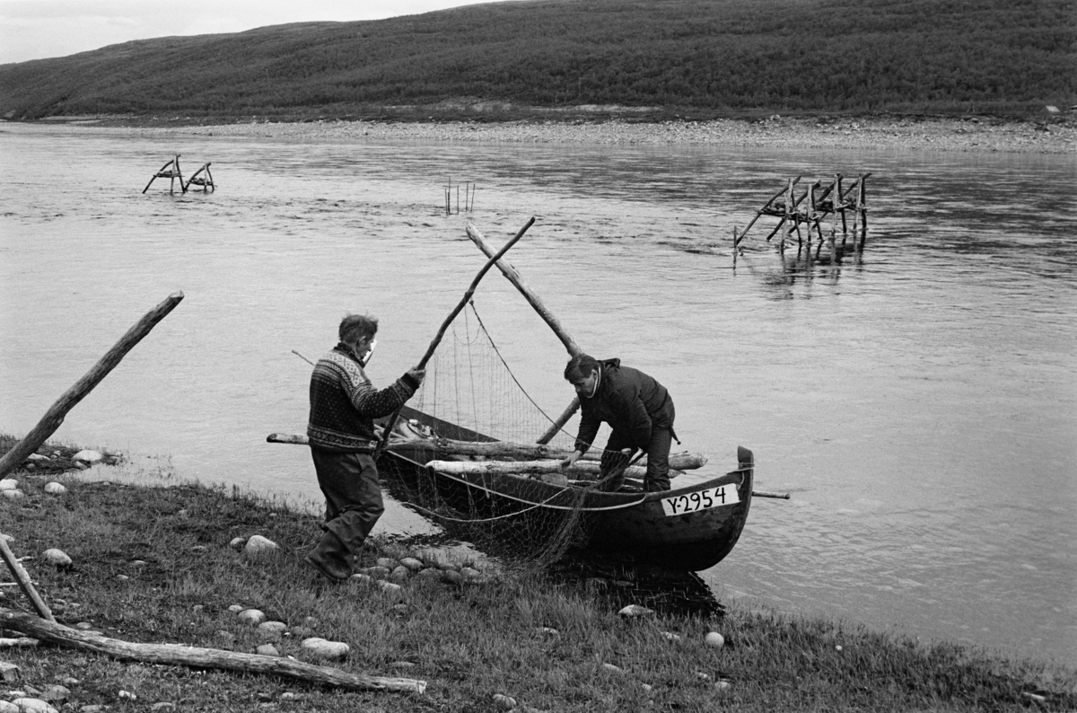 Per (til venstre) og Aslak Varsi legger et posegarn i elvebåten sin, for å kunne montere dette i en stengselsinnretning de har satt opp med sikte på å fange laks i Tana (Deatnu) i Finnmark.  Bildet er tatt litt sør for bygdelaget Sirma.  På vannspeilet i bakgrunnen skimtes en del staker og noen trefotete stativer, som var forankringspunkter for ledegjerder og posegarn som ble brukt under dette fisket.  Et stativ ligger også på tvers av båten, og skal antakelig ros ut for plassering i elva.  Disse trefotingene ble holdt på plass ved hjelp av stein som ble plassert som tyngsel på ei hylle under krysningspunktene i toppen av konstruksjonene.  Slike trefotete stativ ble plassert på steder hvor det ikke lot seg gjøre å drive pæler ned i elvebotnen.  Mellom stakene og trefotingene ble det montert ledegjerder av ris (ukvistet bjørk) eller garnrammer.  Posegarna ble – naturlig nok – montert etter at ledegjerdene var på plass.  Staven Per bar mot båten da dette fotografiet ble tatt ble kalt for goarat.  Den var tredd gjennom maskene i posegarets ytre arm.  Denne fiskemåten – buoððu – ble praktisert i sommersesongen, på lavere vannstand og med roligere strømforhold enn under vårflommen i elva.  Fotografiet er tatt på St. Hansaften i 1975. 