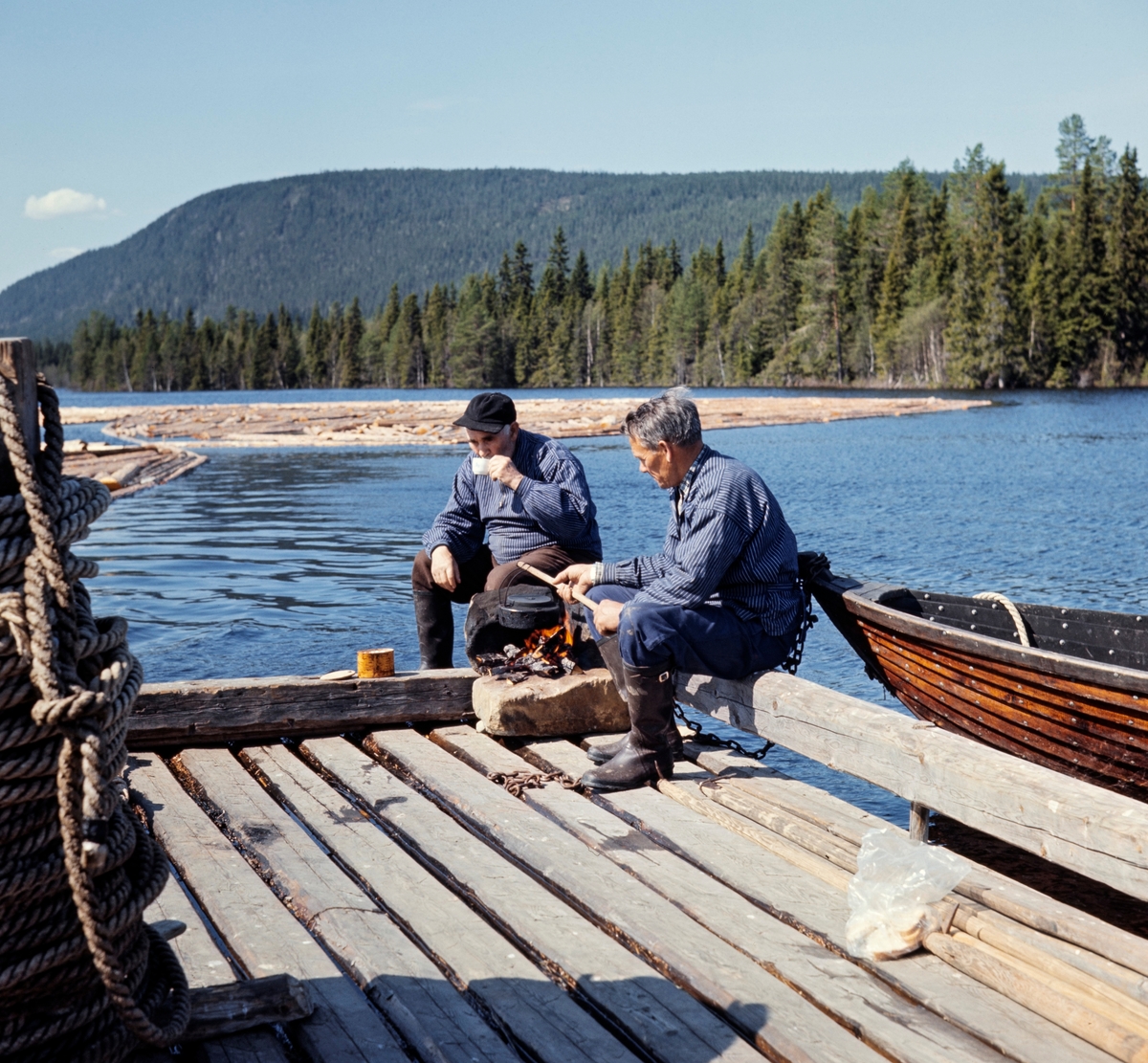 Johan (til venstre) og Halvor Sjølie, fotografert under kaffepause på spillflåte på en av Ulvsjøene i Trysil i slutten av mai 1969.  Karene sitter i et av flåtens fire hjørner, der de hadde gjort seg opp kaffevarme på ei steinhelle.  Halvor holdt kaffekjelen på en kjepp over varmen, mens Johan løftet kaffekoppen til munnen.  Begge karene hadde blå busseruller med kvite striper på overkroppen og gummistøvler på beina.  Johan var kledd i brune vadmelsbukser, Halvor i blå bomullsbukser.  Til venstre i forgrunnen ser vi «tønna» med trossa som ankeret - «krabba» - var festet i.  Bak karene (til høyre) ligger robåten som ble brukt når krabba skulle flyttes.  På vannspeilet i bakgrunnen ligger en tømmerbom.  Ellers viser bildet de skogkledde åsene som preget landskapet omkring Ulvsjøene. 

Litt generell informasjon om spillflåtefløtinga på Ulvsjøene finnes under fanen «Andre opplysninger». Ulvsjøen. Hedmark. 