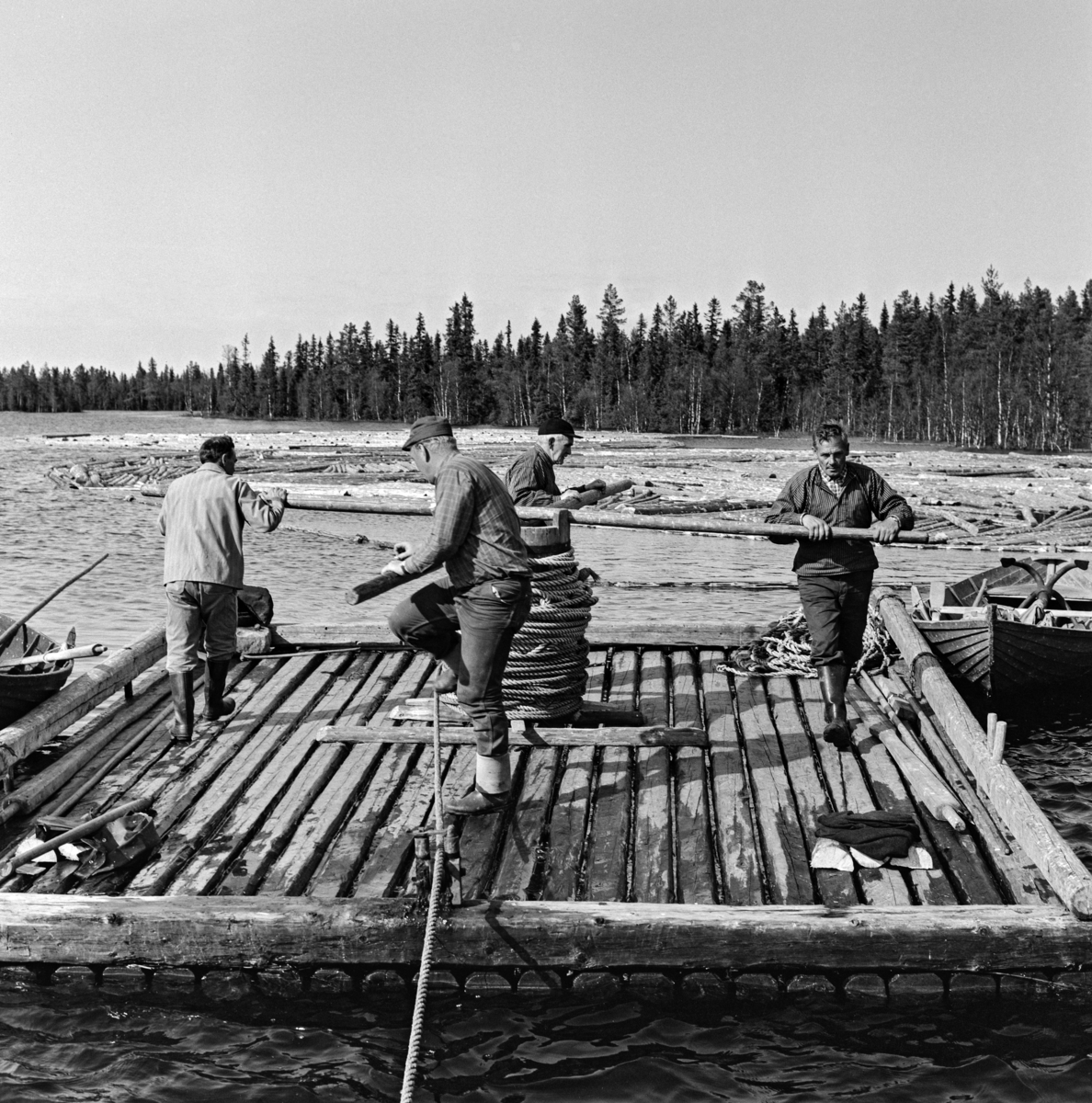 Arbeidslag på spillflåte på en av Ulvsjøene i Trysil i mai 1969.  Fotografiet er tatt mot sentrum av den noenlunde kvadratiske spillflåten, der karene beveget seg på.  I sentrum av denne er det reist en cirka en meter høy sylinder, som ble kalt «tønna».  På toppen av denne roterbare konstruksjonen er det lagt to trestenger - «årer» - i kryss.  Dette er et gangspill med ei cirka 200 meter lang trosse med et anker - kalt «krabba» - i ytterenden.  Krabba ble rodd framover i sjøen, så langt trossa rakk, og sluppet ned på botnen, hvor den forhåpentligvis festet seg.  Deretter kunne karene «spille» - dreie gangspillet rundt og rundt - slik at flåten og en bakenforliggende tømmerbom sakte men sikkert ble trukket mot ankringspunktet.  Når de nådde fram dit, ble krabba hevet og rodd videreframoveri den retningen man ønsket å ha tømmereet.  En av robåtene som ble brukt ved slike forflytninger skimtes til venstre i bildet.  Fotografiet illustrerer hvordan mannskapet på flåten måtte løfte beina høyt for å unngå snubling hver gang de passerte trossa. Arbeidslaget på fotografiet skal være (fra venstre) Torfinn Bakken (med ryggen til), Magne Neby, Johan Sjølie og Halvor Sjølie.  

Litt generell informasjon om spillflåtefløtinga på Ulvsjøene finnes under fanen «Andre opplysninger». Ulvsjøen. Hedmark.