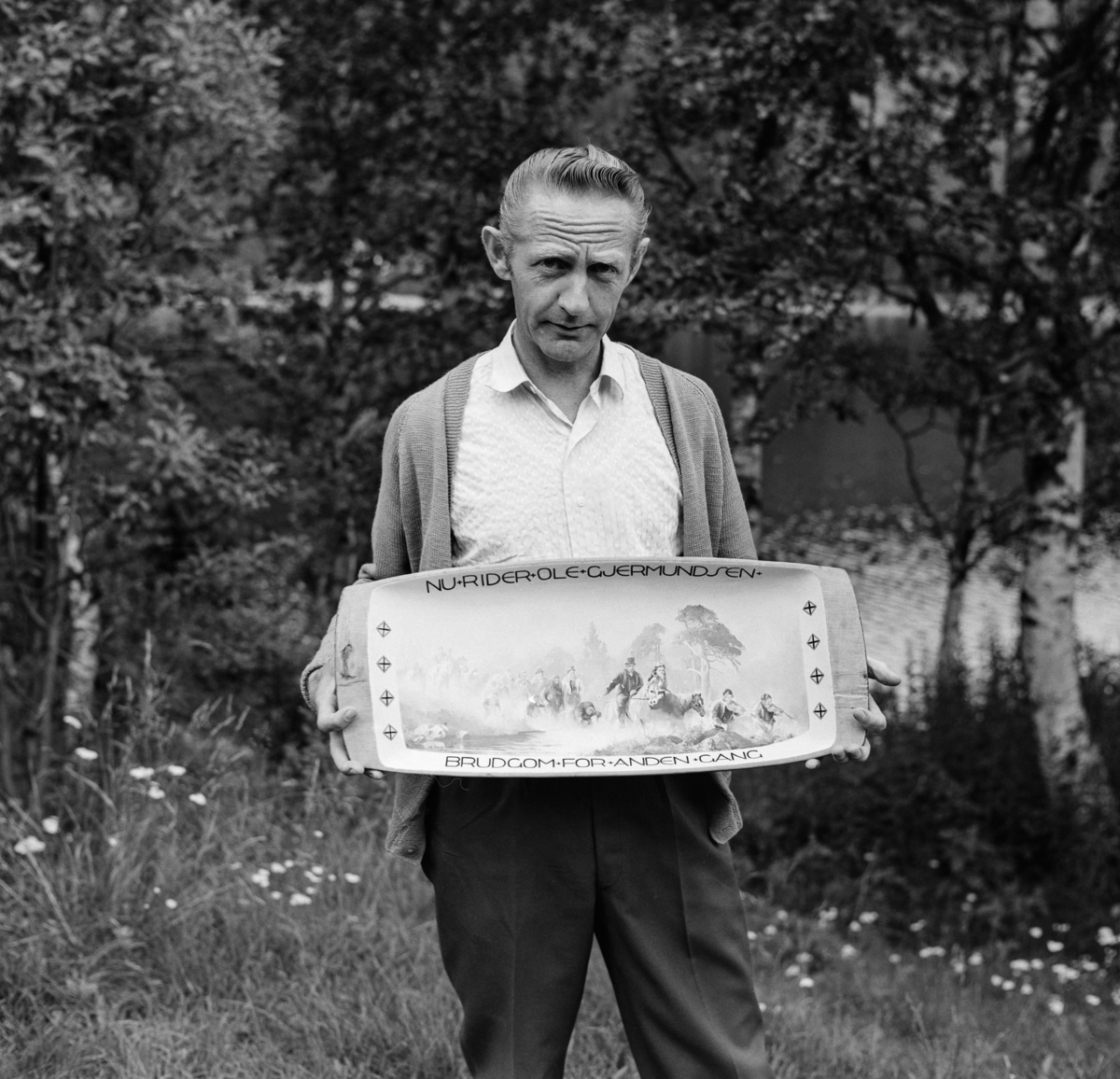 Maleren Gunvald Sand fra Engerdal, fotografert i 1967.  Bildet er tatt utendørs.  Sand poserer med et tretrau, som han har dekorert innvendig med ei framstilling av et ridende brudefølge i et landskap som kan som kan minne om engerdalsnaturen.  Over og under bildet har han skrevet følgende tekst: «NU RIDER OLE GJERMUNDSEN BRUDGOM FOR ANDEN GANG». 