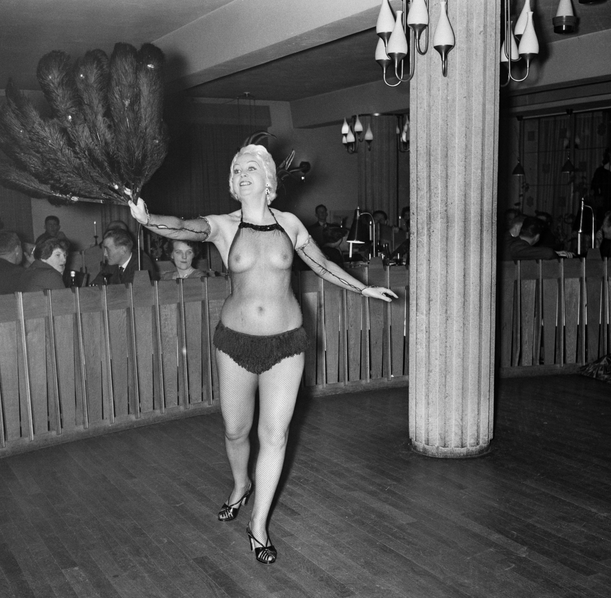 Fra stripteaseforestilling på restaurant Elvarheim i Storgata i Leiret (Elverum sentrum) tidlig i 1960-åra.  Fotografiet viser ei lyshåret kvinne på det parkettbelagte dansegulvet som var skjermet mot de omkringstående bordene, hvor gjestene spiste og drakk.  Danseren hadde sko og ei truse på seg.  Armene, overkroppen og beina var iført et grovmasket nettingstoff, som på ingen måte skjulte kvinnekroppen.  I høyre hånd holdt aktøren ei diger fjærvifte, som antakelig hadde vært brukt til å dekke «pirrende» kroppsdeler i den erotisk ladde forestillingens innledende fase.  Musikken til forestillingen ble framført at tre mannlige restaurantmusikere, som spiller klaver, kontrabass og trommer (jfr. SJF-F. 006996).  Nakenhet var tabubelagt da dette fotografiet ble tatt, og følgelig ganske eksotisk i et litet lokalsamfunn som Elverum.  Restauranten lå i underetasjen av den fløyen av kommunens rådhus som vendte mot Storgata.  Bygningen var tegnet av arkitektfirmaet Torp & Torp, og sto ferdig i 1956.  Restaurant Elvarheim hadde derfor svært moderne lokaler da dette bildet ble tatt. 