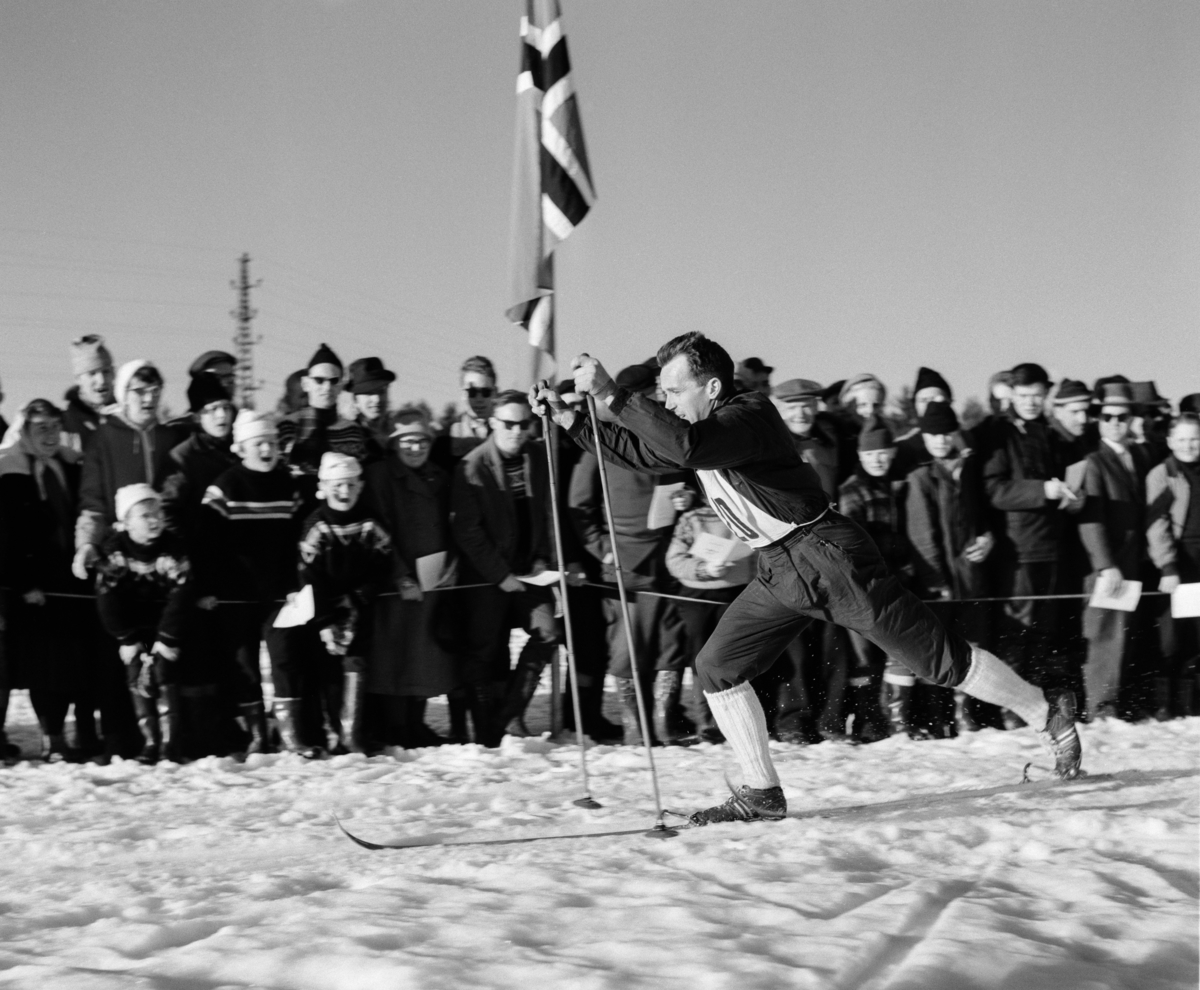 Langrennsløperen Hallgeir Brenden (1929-2007), fotografert i et skirenn han deltok i vinteren 1961.  Bildet er tatt på ei flate der Brenden gikk med dobbelttak – han lente kroppen forover, satte stavene i snøen like framfor høyrefoten og skjøv fra samtidig som han sparket til med den nevnte foten.  Dermed skled han noen meter før han gjentok dette bevegelsesmønsteret.  Brenden var kledd i nikkersbukser med kvite strømper.  Han gikk på treski med «Rottefella-bindinger» og hadde bambusstaver.  At han verken hadde votter eller lue kan vel tyde på at dette fotografiet er fra et renn som gikk under forholdsvis moderate temperaturforhold mot slutten av sesongen.  Løperen heies fram av entusisastiske tilskuerne, som står tett bak ei tausperring.  Hallgeir Brenden var fra Tørberget i Trysil.  Han representerte Vestre Trysil IF, og var langrennsløper av internasjonalt format i 1950-åra.  Høydepunktene i karrieren var olympiske seiere på 18 kilometer i 1952 og på 15 kilometer i 1956, men han var også en av løperne på det norske stafettlaget som vant olympisk sølv i 1960.  Brenden var dessuten friidrettsutøver for IK Tjalve.  Han var i norgeseliten i lange baneløp, men oppnådde ingen internasjonale triumfer med piggsko på beina. 