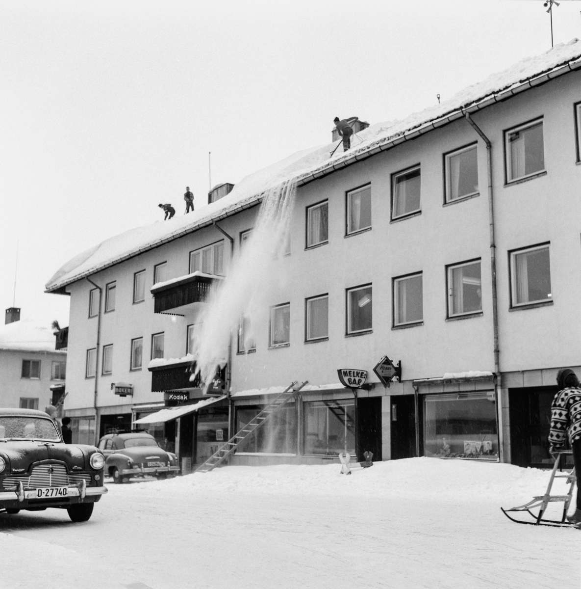 Snøen måkes fra taket på Skogbrukets hus i St. Olavs gate i Elverum.  Vi skimter tre karer med snøskuffer på taket, hvor det lå et tjukt og antakelig tungt snølag på det forholdsvis slake saltaket.  Snøen kastes ned på fortauet, som var midlertidig sperret for fotgjengere ved hjelp av en stige.  Dette, og moderat biltrafikk, var antakelig årsaken til at ei kvinne med spark ved høyre bildekant holder seg i vegbanen.  Skogbrukets hus var en solid treetasjes bygning med forretninger i lokalene i første etasje mot St. Olavs gate (bokhandel, fotoforretning og serveringsstedet «Melkebaren») og med kontorer og leiligheter i etasjene over.  Skogetatene for Elverum kommune og Hedmark fylke var blant dem som hadde kontorer i bygningen.  Bilen ved venstre bildekant er en Ford Sephyr Six.  Årsmodellene fra perioden 1951-1956 hadde slik front.

Det var daværene Elverum-ordfører Otto Ødegård som tok initiativet til å få bygd et «Skogbrukets hus» i Elverum, blant annet for å kunne samle den kommunale og den fylkeskommunale skogadministrasjonen under ett tak.  I 1949 ble det oppnevnt en komité som skulle arbeide med saken.  Komiteen tok kontakt med arkitekt Otto L. Scheen i Oslo, som tegnet et forretnings- og kontorbygning i funkisstil.  Bygginga av huset ble organisert gjennom et aksjeselskap, der Elverum kommune bidro med 100 000 kroner.  Det lokale entreprenørselskapet Martin M. Bakken fikk byggeoppdraget.  Bygningen sto ferdig i 1953 og kostet om lag 700 000 kroner.  Ferdigstillelsen skjedde altså det samme året som den store, nasjonale skogutstillingen ble arrangert i Elverum.