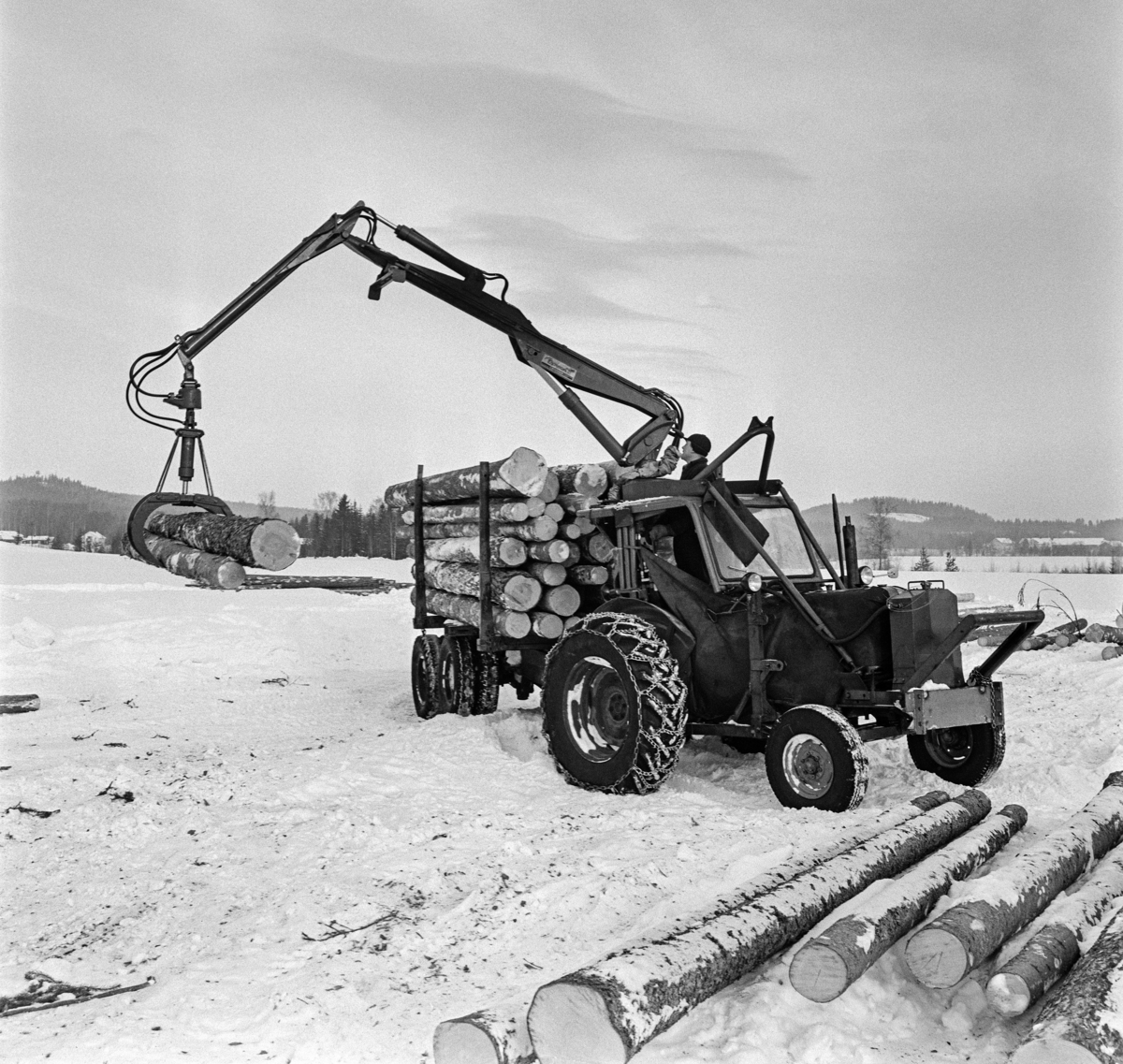 Tømmerkjøring med traktor, muligens på Jømna i Elverum kommune i Hedmark vinteren 1969.  Traktorer med tømmertilhenger og griplasterkran ble lansert som en nyhet i slutten av 1960-åra.  Da hadde den svenske produsenten HIAB lansert sin «skogselefant».  De norske skogteknologene var skeptiske til denne maskintypen.  Med den norske eiendomsstrukturen og i norsk terreng var det de rammestyrte helstammelunnerne som var framtidsrettet, hevdet de.  Mjøsen, Glommen og Nedre Glommen skogeierforeninger satte likevel i gang forsøk med det de kalte «kombinasjonsmaskiner» - traktorer med griplastere og boggidoninger.  I slutten av 1960-åra kunne norske maskinleverandører tilby flere varianter av slike maskiner.  Den svenaske «skogselefanten» med HIAB-kran på en rammestyrt traktor var den mest avanserte, men det ble også levert svenske og finske krantyper som kunne monteres på kraftige landbrukstraktorer.  Også den norske maskinfabrikken Hymas i Brumunddal bygde tidlig ei gripeklo som kunne monteres i steden for skuffa på sine traktorgravere, som allerede var mye brukt i det norske skogbruket.  Dermed kunne man få helårsdrift på en traktor som inntil da i hovedsak hadde vært brukt sommerstid.  Som tømmerlastingsmaskin var Nuffieldene med kraner fra Hymas mest anvendelige på velteplasser, men de kunne også trekke tilhengere med tømmer som var pålesset ved hjelp av traktorkrana.  Det er usikkert om det vi ser på fotografiet er et Hymas-produkt, eller om det er en svensk modell.  

Etter hvert måtte de rammestyrte stammelunnerne vike plassen for «kombinasjonsmaskiner» med kran og tilhenger for tømmerlass.  Skogteknologene gav dem etter hvert betegnelsen «lastetraktorer», mens maskinforhandlerne brukte betegnelsen «lassbærere», for å tydeliggjøre at det tømmeret man fikk fram ikke ville være sølete og tilgriset, slik stammelunnet virke kunne være, i hvert fall i sommerhalvåret.