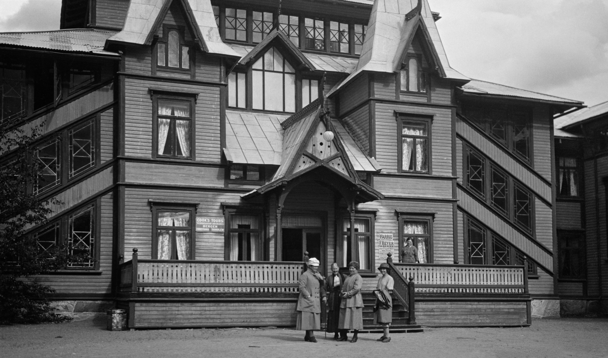 Fire kvinner foran trappa som førte inn i Dalen hotell.  Hotellet ligger, som navnet antyder, i tettstedet Dalen i Tokke kommune i Telemark.  Etter at Bandak-Norsjø-kanalen ble åpnet i 1892 ble Vest-Telemark et turistmål.  Turistene kom på dampbåtene, som gikk mellom Skien og Dalen.  Ved rutas endepunkt ved enden av innsjøen Bandak var det ikke noe overnattingssted som velstående reisende fant attraktivt før dette hotellet sto ferdig i 1894.  De ble bygd etter tegninger av arkitekten Haldor Larsen Børve, som gav bygningen et storslått og nasjonalt preg, med tårn, spir, drakehoder, detaljrike altaner, profilerte rekkverk og paneler.  Hotellet har et areal på bortimot 4 500 kvadratmeter, og hadde mange celebre gjester de første åra det var i drift.  Under 2. verdenskrig overto tyske okkupasjonsstyrker bygningen, og deres bruk av lokalene var neppe særlig pietetsfull.  I åra som fulgte ble forfallet gradvis tydeligere.  Predikanten Aage Samuelsen kjøpte det etter hvert med sikte på å få det til å gjenoppstå i sin gamle prakt, men greide ikke å reise midler til det.  Han fikk imidlertid åpnet andres øyne for dette helt spesielle kulturminnet.  Hotellet ble gjenåpnet i 1992.  Dette fotografiet ble tatt 70 år tidligere, nærmere bestemt 18. juni 1922. 