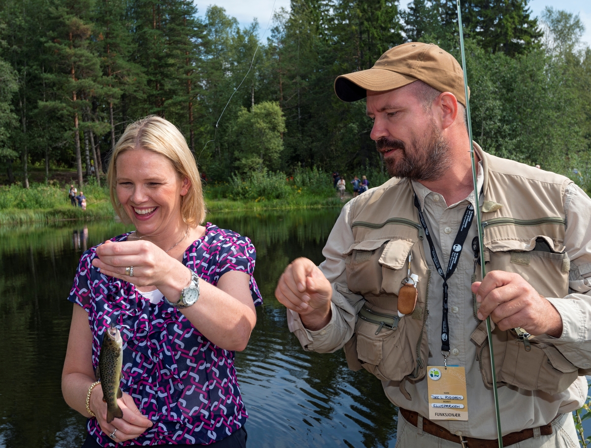 Landbruks- og matminister Sylvi Listhaug og fiskeinstruktør Tore Litleré Rydgren fotografert ved Norsk Skogmuseums fiskedam på Prestøya i Glomma under De nordiske jakt- og fiskedager torsdag 7. august 2014.  Rydgren gav politikergjesten et lite fiskekurs fra en plattform ved dammen, og Listhaug fanget en liten ørret, som hun løftet i brysthøyde for å se på da dette fotografiet ble tatt.  Fisken ble kort tid etterpå sluppet tilbake i vannet.  Landbruks- og matministeren besøkte jakt- og fiskedagene som foredragsholder i seminaret «Gull og grønne skoger – nye grep for å øke skogens verdi og glede for alle» i museets auditorium.  Etterpå benyttet hun sjansen til å oppleve noe av programmet i museets friluftsmuseum, blant annet her ved fiskedammen. 