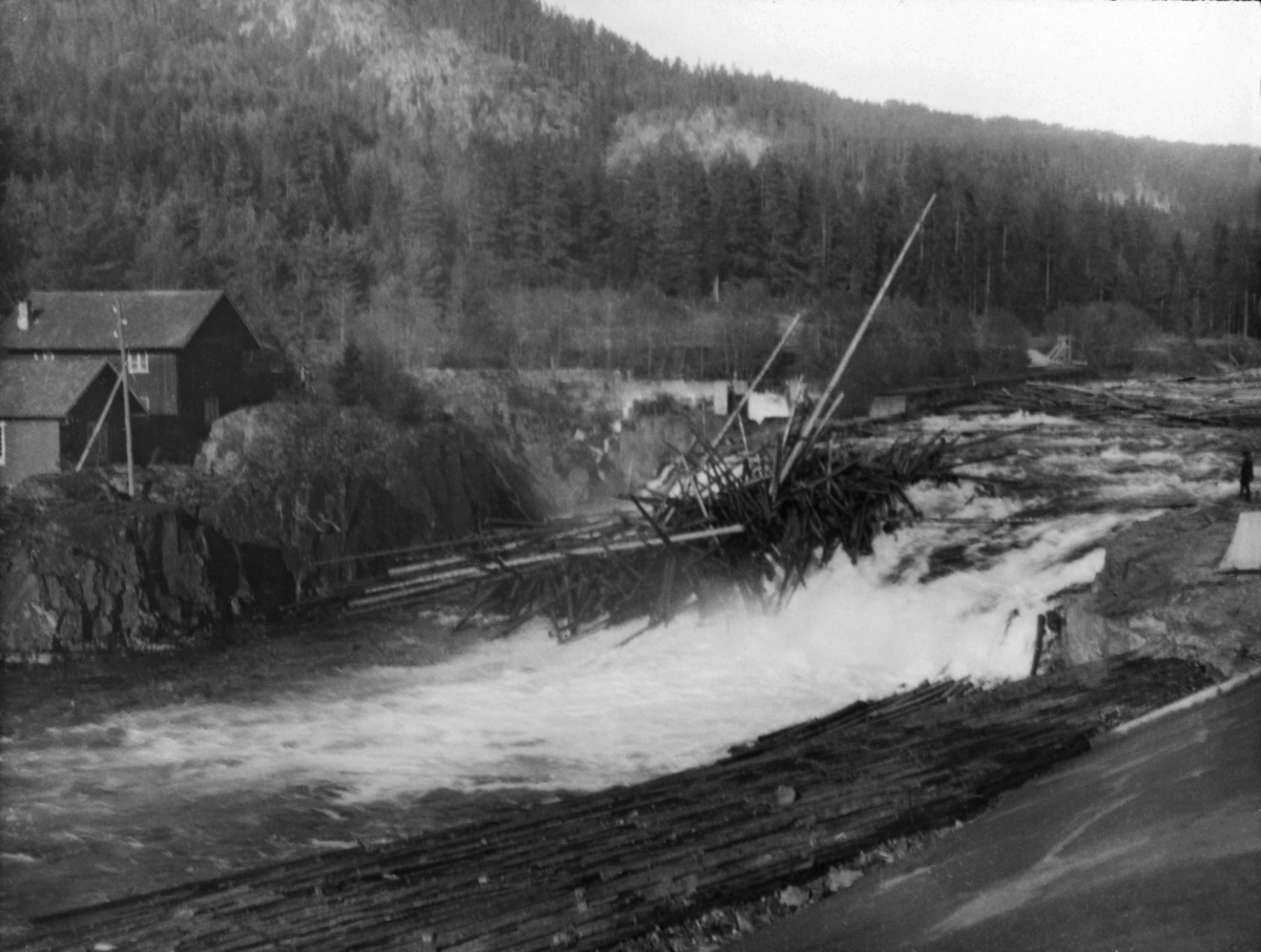 Tømmerhaug i Sagafoss i Tinnåa (Tinne) i Notodden (tidligere Heddal eller «Hiterdal») kommune i Telemark.  Fotografiet er tatt i motstrøms retning fra østre elvebredd, mot et kvitskummende, flomstort fossefall med fløtingstømmer som har satt seg fast i hauger eller vaser, spesielt langs elvebreddene.  På motsatt side av elva derimot, på vestsida av fossen, ligger stokkene hulter til bulter, sprikende i mange retninger.  Også i strykene ovenfor det bratteste fallet skimter vi tømmervaser.  Vest for fossen lå det et halvannenetasjes våningshus med tilhørende uthus.  I bakgrunnen ser vi den skogkledde Tinneåsen.  Sagafossen var om lag ti meter høy, og den voldte atskillige ulemper for fløtinga i Tinnåa, for det hadde lett for å sette seg tømmerhauger under dette vannfallet, både når det var høy og når det var lav vannføring.  I slutten av 1890-åra ble det bygd tømmerskjermer på begge sider av fossen, noe som skal ha redusert problemet.  Disse skjermene er imidlertid ikke å se på dette fotografiet.  Sagafossen ble helt borte da den nye Tinnfossdammen ble tatt i bruk i midten av 1950-åra. 