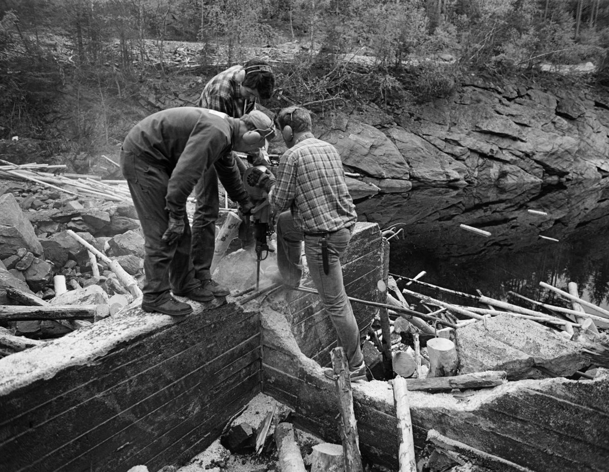 Fjerning av damløp ved Osfallet kraftstasjon i elva Søndre Osa i Åmot i Hedmark sommeren 1984.  Dette var den siste sesongen det ble fløtet i denne delen av Glomma-vassdraget, og ettersom dette var ei planlagt og definitiv avvikling av virksomheten, pålå det Glomma fellesfløtningsforening å fjerne installasjoner i vassdraget som på kort eller lang sikt kunne representere en fare eller være til hinder for annen virksomhet.  I dette tilfellet er det grunn til å anta at tømmerløpet ble revet i samarbeid med selskapet som eide og drev kraftanlegget.  På fotografiet er vi tre karer i arbeid med en bormaskin som ble brukt til å angripe et fundament av armert betong.  På dette underlaget hadde det ligget et golv av trebjelker, som skulle hindre at fløtingstømmeret hektet seg fast i den ujamne bergflata på elvebotnen i tømmerløpet.  Nedenfor arbeidsstedet ser vi en del tømmer som er kappet i korte kabber samt et elveparti der det var uvanlig liten vannføring, antakelig fordi dammen var stengt slik at demonteringsarbeidet kunne skje uten at vannet ble noe hinder.  Dammen på fotografiet skal ha vært ferdigstilt i 1947.  Den ble noe ombygd i 1981. Energiproduksjonen foregår i en kraftstasjonsbygning fra 1914, hvor det er installert to francisturbiner som kan yte 5.5 megawatt.  Den opprinnelige dammen ble ødelagt ved et dambrudd i 1916.  Det nåværende kraftverket utnytter et fall på 41 meter i elva Søndre Osa.