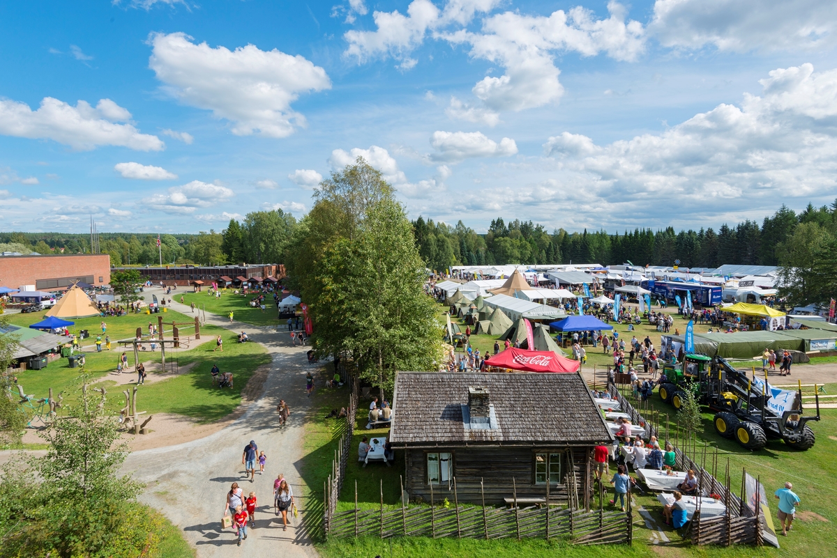 Utsikt fra balkongen utenfor oppholdsrommet i det rekonstruerte brannvakttårnet på Norsk Skogmuseum i Elverum.  Fotografiet er tatt mot gangvegen mellom museumsbygningen og friluftsmuseet på Prestøya søndag 10. august 2014, den siste dagen under De nordiske jakt- og fiskedager dette året, noe som betydde at det var flere mennesker og mer aktivitet på uteområdet enn på en vanlig søndag.  Til høyre i forgrunnen ser vi skogvokterboligen som er flyttet til museet fra Galten i Engerdal.  Ved dette huset var det serveringssted med salg av kaffe, vafler og is under arrangementet.  Til venstre i forgrunnen ser vi utkanten av lekeområdet for barn o bakenfor ser vi lavvoen der den statlige skogforvaltningen presenterte sin virksomhet for publikum.  På motsatt side av gangvegen ser vi det såkalte «håndverkstorget», til venstre for trerekka som står i grensesonen mellom det som for noen tiår siden var skillet mellom to åkrer. På sørsida av denne ser vi det såkalte «frilandsområdet», der de fleste av arrangementets kommersielle aktører var samlet.  Lassbæreren til høyre i forgrunnen står der som et landemerke som markerer det såkalte «skogbrukstunet» på bakkekammen bak våningshuset på klokkergarden Fossum, der organisasjonene i skogbruket hadde sine aktiviteter under arrangementet.  Den dagen da dette fotografiet ble tatt var det 6 789 personer som løste billetter til De nordiske jakt- og fiskedager.