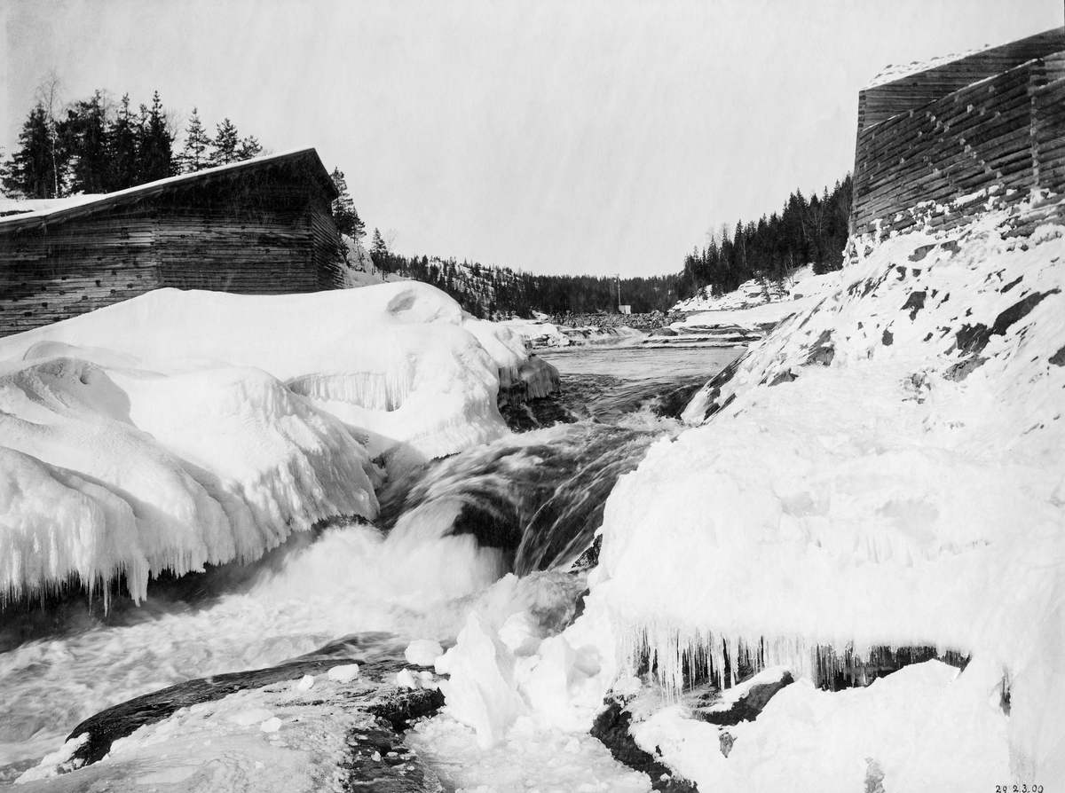 Fossefall, antakelig ved Kykkelsrud, der elva er grenselinje mellom Spydeberg kommune i vest og Askim i øst.  Bildet er tatt vinterstid, i motstrøms retning, ved et lite fossefall der det på dette tidspunktet var bare midtålen som var åpen, mens det lå is på begge sider.  Ovenfor fossen ser vi steinfylte tømmerkistekar ved begge elvebredder, antakelig bygd med sikte på å styre strømretningen og tømmerets gang i det nedenforliggende elveløpet ved flomvannstand.  Ovenfor skimter vi en molo av naturstein.  Den ble antakelig bygd i samband med at A/S Glommens Træsliberi i perioden 1899-1903 bygde kraftstasjon som hentet vann fra e dam ved Strandefossen ovenfor og en kraftstasjon ved Rudsfossen (Hvervefossen) om lag en kilometer lengre nede, en fallhøyde på 19 meter.  Fotografiet er antakelig tatt som dokumentasjonsgrunnlag med sikte på forhandlingene mellom Fredrikstad Tømmerdirektion og kraftutbyggerne om hva som skulle gjøres i elveløpet for sikre at det – med den reduserte vannføringa kraftproduksjonen innebar – fortsatt kunne fungere som transportled for fløtingstømmer.