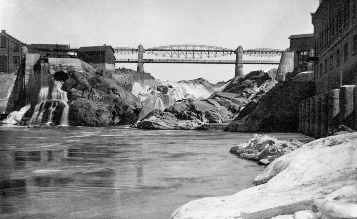 Sarpsfossen eller Sarpefossen i Glomma, fotografert i motstrøms retning i en situasjon da det var ekstremt liten vannføring i elva.  Bildet er tatt fra ei bergflate ved elvebredden på østsida av vassdraget.  Vi ser en teglsteinsmurt industribygning på «Hafslund-sida» til høyre i bildet, og tilsvarende bebyggelse på «Borregaard-sida» til venstre.  I midten ser vi sjølve fosseløpet, der de på dette tidspunktet sparsomme vannmassene framstår som et kvitt slør over de bergknausene som tross alt var dekt av vann.  Over det hele rager den staselige veg- og jernbanebrua som ble bygd like ovenfor fossen i 1870-åra mot himmelen.  Fotografiet antas å være tatt omkring år 1900.