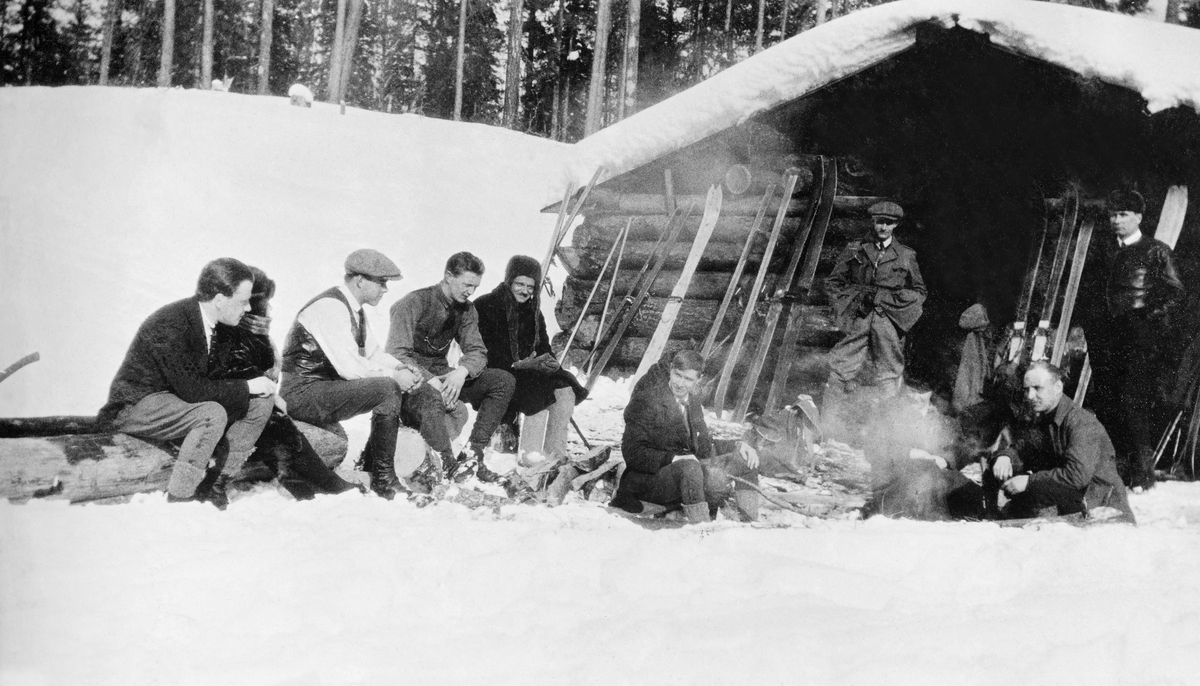 Skandinavene som drev sagbruk i byene ved utløpene av de store nordvestrussiske elvene tidlig på 1900-tallet holdt sammen, blant annet ved å gå skiturer sammen på vintersøndager.  Her ser vi sju menn, samlet omkring et bål foran ei laftet tømmerkoie.  Karene hadde stilt skiene sine opp mot koieveggen mens de slappet av og snakket sammen.