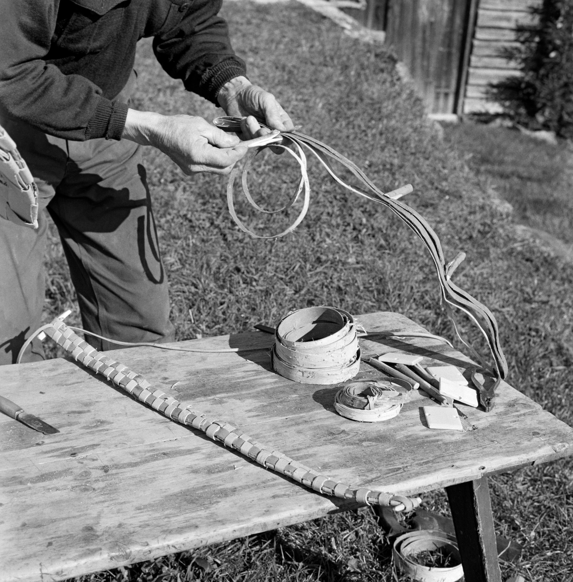 August Herstadhagen (1886-1977) fra Elverum demonstrerer kontfletting på en benk han hadde stilt opp i tunet på gardsbruket sitt.  Konten flettes av en til halvannen tomme brede neverstrimler.  Da dette fotografiet ble tatt hadde Herstadhagen gjort seg ferdig med sjølve konten.  Han arbeidet med feslingene – skulderstroppene redskapet skulle bæres i.  En av dem var ferdigflettet og lå på benken, utstyrt med endepinner.  Disse skulle forankres i små treplater inne i neverkonten, for flettverket var ikke kraftig nok til at det kunne tjenes som festepunkt.  I hendene holdt Herstadhagen neverstrimler som antakelig skulle brukes i den andre ferslingen.  Disse ble lagt oppå hverandre og midlertidig sammenføyd ved hjelp av små seljepinner som var splittet i den ene enden, slik at de kunne fungere som klyper.