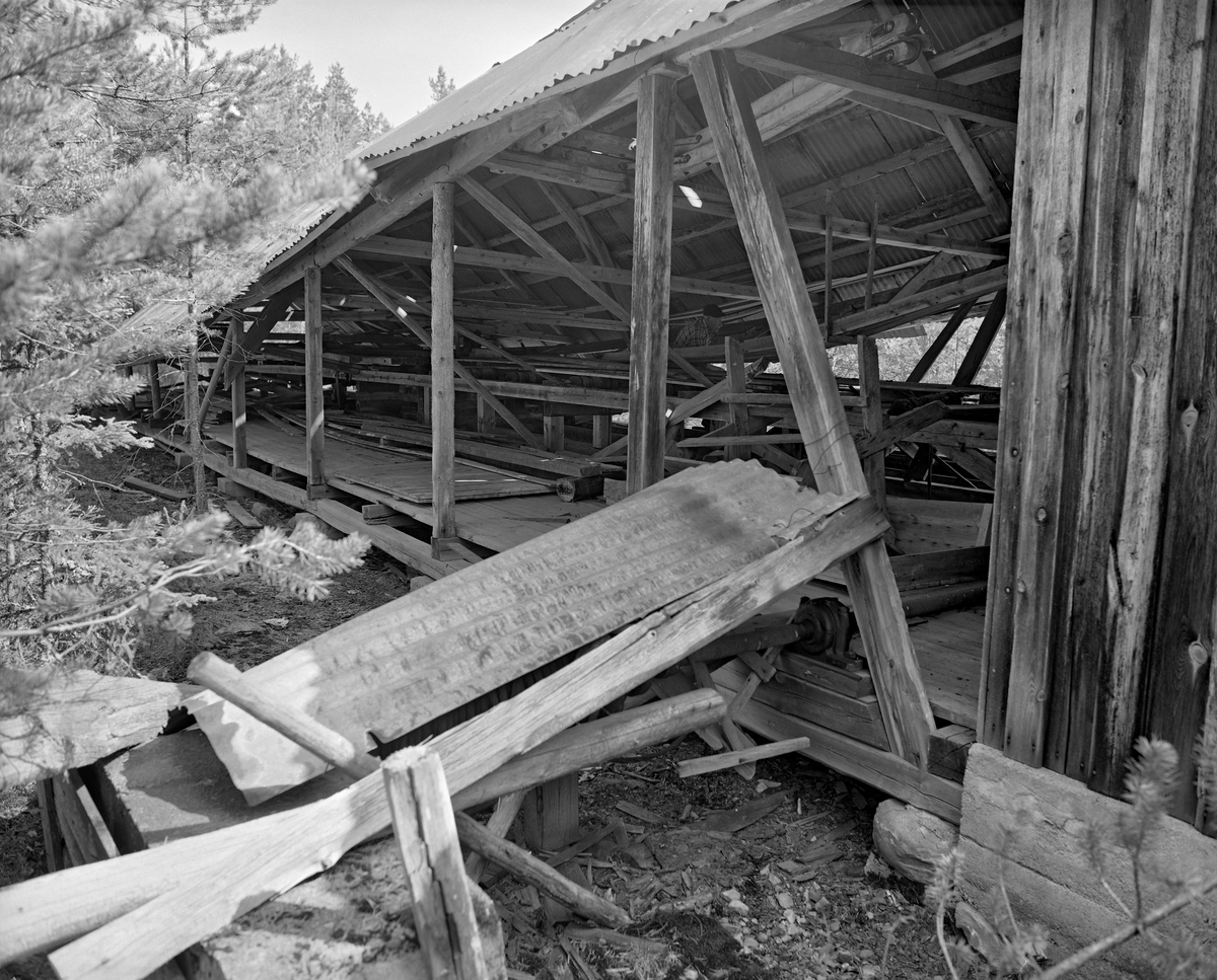 Eksteriør fra Mastveltsaga i Åsnes.  Fotografiet er sommeren 1975, da lokomobilen som drev saga og den øvrige innredningen ble demontert og flyttet til et nytt saghus på Norsk Skogbruksmuseum (Norsk Skogmuseum) i Elverum.  Saghuset på Mastvelta var en bindingsverkskonstruksjon, som i den ene enden var kledd med over- og underliggende panel av vankantete bord, mens sagbenken sto i den delen av bygningen som ikke hadde bordkledde vegger.  Dette fotografiet er tatt mot den ene langveggen, tett ved overgangen mellom den bordkledde og den uinnkledde delen av bygningen.  I forgrunnen ser vi ei bølgeblikkplate som lå som overdekning over en jernspole som det var viklet en del vaier på.  Denne innretningen (en slags kjerrat) ble antakelig brukt til å trekke stokker fram mot saghuset og sagbenken, som vi skimter inne i bygningen. Trongaardsaga. Lokomobilsag.

Mer informasjon om Mastveltsaga og om lokomobilsager generelt finnes under fanen «Opplysninger».