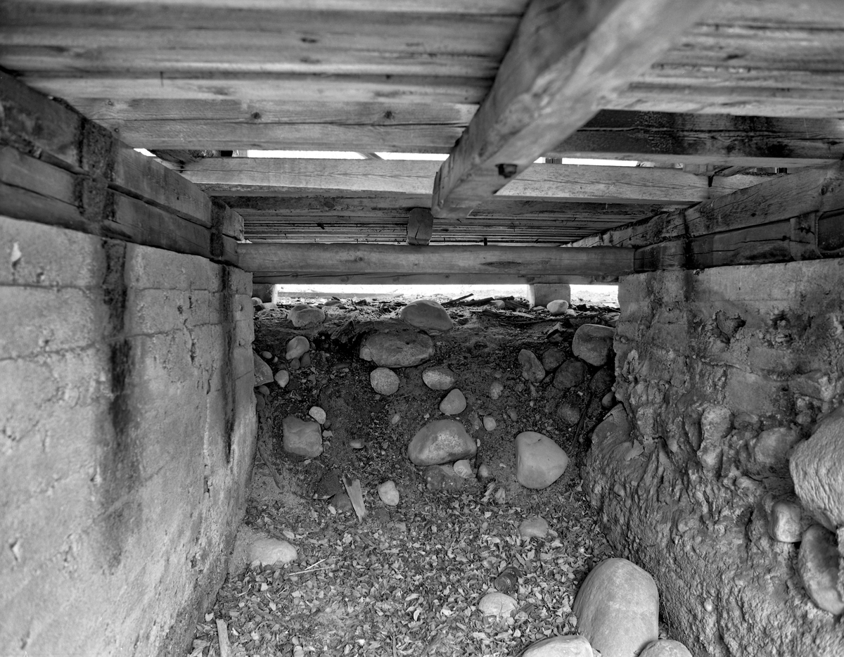 Fra underbygningen under saghuset på Mastveltsaga i Åsnes i Solør.  Fotografiet er sommeren 1975, da lokomobilen og saginnredningen fra dette anlegget ble demontert og flyttet til et nytt saghus på Norsk Skogmuseum (fra 2003 Norsk Skogbruksmuseum) i Elverum.  Dette fotografiet viser hvordan en del av bygningen var reist av firskårne bjelker på støpte undermurer.  Dette gjaldt for øvrig bare deler av bygningen, mye av saghuset kvilte også på støpte pilarer og laftestein.  På det stedet der dette fotografiet ble tatt var det imidlertid en nedgravning i bakken mellom to støpte vegger - antakelig et flisrom som var slik innrettet at det stundom kunne tømmes.  Flis fra denne saga ble for øvrig brukt i et eget brennkammer som gav varmeenergi til lokomobilen (jfr. SJF-F.008930).

Mer informasjon om Mastveltsaga og om lokomobilsager generelt finnes under fanen «Opplysninger».