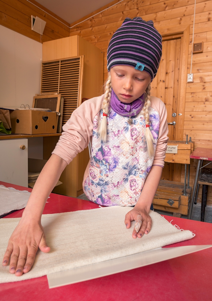 Fra det såkalte «papirkjøkkenet» ved Klevfos Industrimuseum på Ådalsbruk i Løten, Hedmark.  Fotografiet viser ei jente fra 2. trinn ved Stavsberg skole i Furnes (Ringsaker kommune), som bretter filt rundt et vått flak med papirmasse, som hun nettopp hadde hentet opp fra et kar ved hjelp av ei finmasket nettingramme.  Papirarkene skal deretter settes i press.  Ved Klevfos Industrimuseum håndlages papir på denne måten som en introduksjon til hvordan papir framstilles i treforedlingsindustriens maskiner.  Prosessen med filtomslagene presenteres som en parallell til den prosessen som foregår i papirmaskinens pressparti.