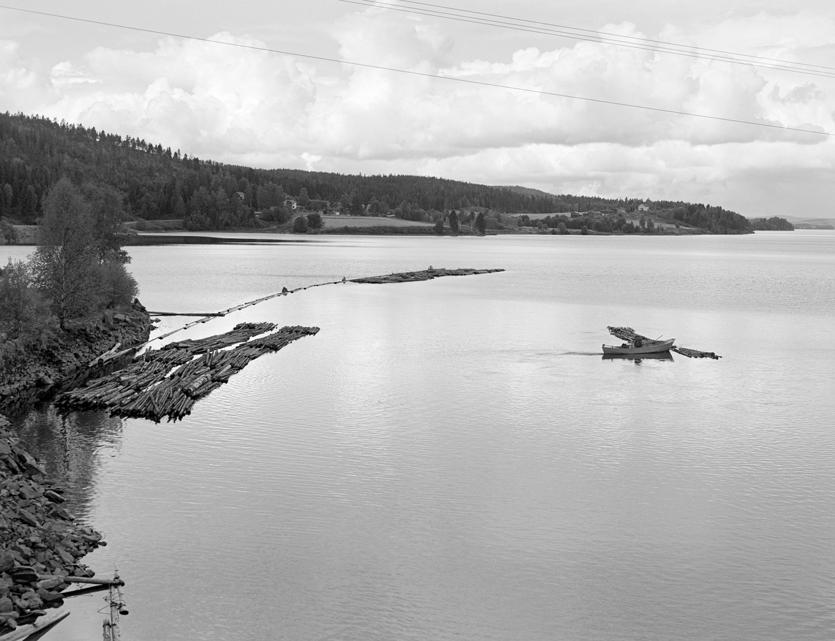 Fra vestre side av innløpet til Ørje sluser i Marker kommune i Østfold (Haldenvassdraget).  Dette sluseanlegget hadde tre kamre, som utliknet en høydeforskjell på ti høydemeter mellom Rødenessjøen (på bildet) og den nedenforliggende Ørjeelva.  Dette fotografiet ble tatt høsten 1982, som var siste fløtingssesong i dette vassdraget.  Da ble det utelukkende fløtet ubarket massevirke (papirråstoff) i tre meters lengder.  Tømmer fra skogene lengst nord i Østfold og sør i Akershus og Hedmark ble utislått fra lastebiler ved Skulerud i Høland og slept ned til Ørje.  Der måtte slepene deles i kortere lengder, som fikk plass i slusekamrene.  Med denne typen tømmer gikk det vanligvis fire bunter – «moser» – i hver slusevending.  Da dette fotografiet ble tatt var fløteren Einar Melby i ferd med å buksere ei slik slusevending mot slusene med en varpebåt av stål. Andre slusevendinger lå fortøyd langs lensa, der lensestokkene var lagt med overlappende ender i medstrøms retning (som takstein), slik at mosene ikke skulle hekte seg fast. 

Det første sluseanlegget ved Ørje ble bygd i perioden 1857-1860, og fra 1877 ble tømmer fra de øvre delene av vassdraget fløtet ned til industrien i Halden på et kanalisert vassdrag.  En kort historikk om fløtinga i Haldenvassdraget finnes under fanen «Opplysninger».