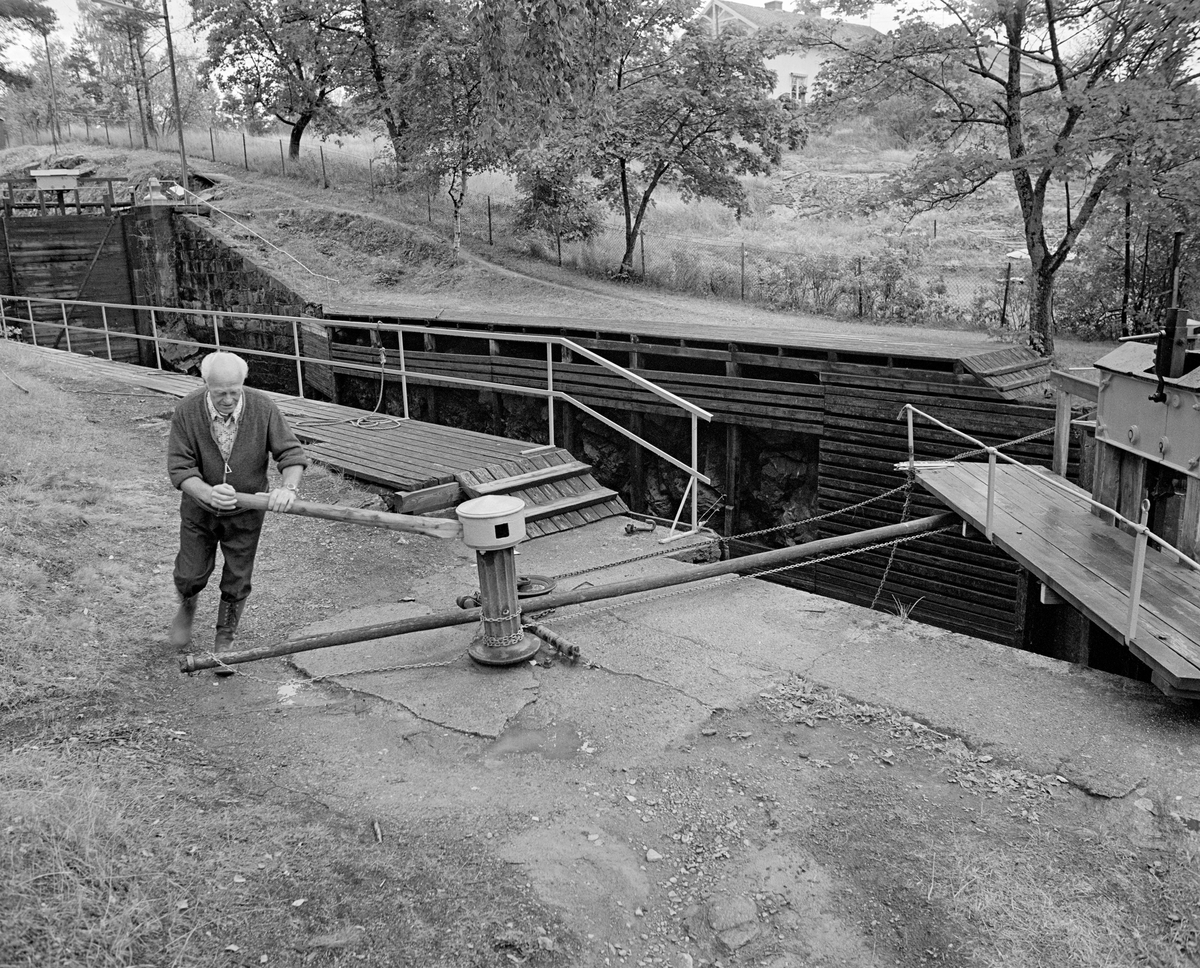 Slusemester Odd Johansen (1917-1993) stenger en av sluseportene ved Ørje sluser i Marker i Østfold.  Dette gjøres ved hjelp av et gangspill med en kjetting og ei lang, kraftig stang som fungerer som skyvearm.  Slusene ved Ørje har tre kamre, og dermed fire porter, som alle manøvreres på denne måten.  Odd Johansen var i mange år slusemester ved nedenforliggende Strømsfoss sluser.  Da dette fotografiet ble tatt, i 1982, var han imidlertid pensjonist, og hadde dermed mulighet for å hjelpe til også på Ørje.  Dette var den siste fløtingssesongen i Haldenvassdraget.

En liten historikk om tømmerfløting og kanaliseringsarbeid i Haldenvassdraget finnes under fanen «Opplysninger».