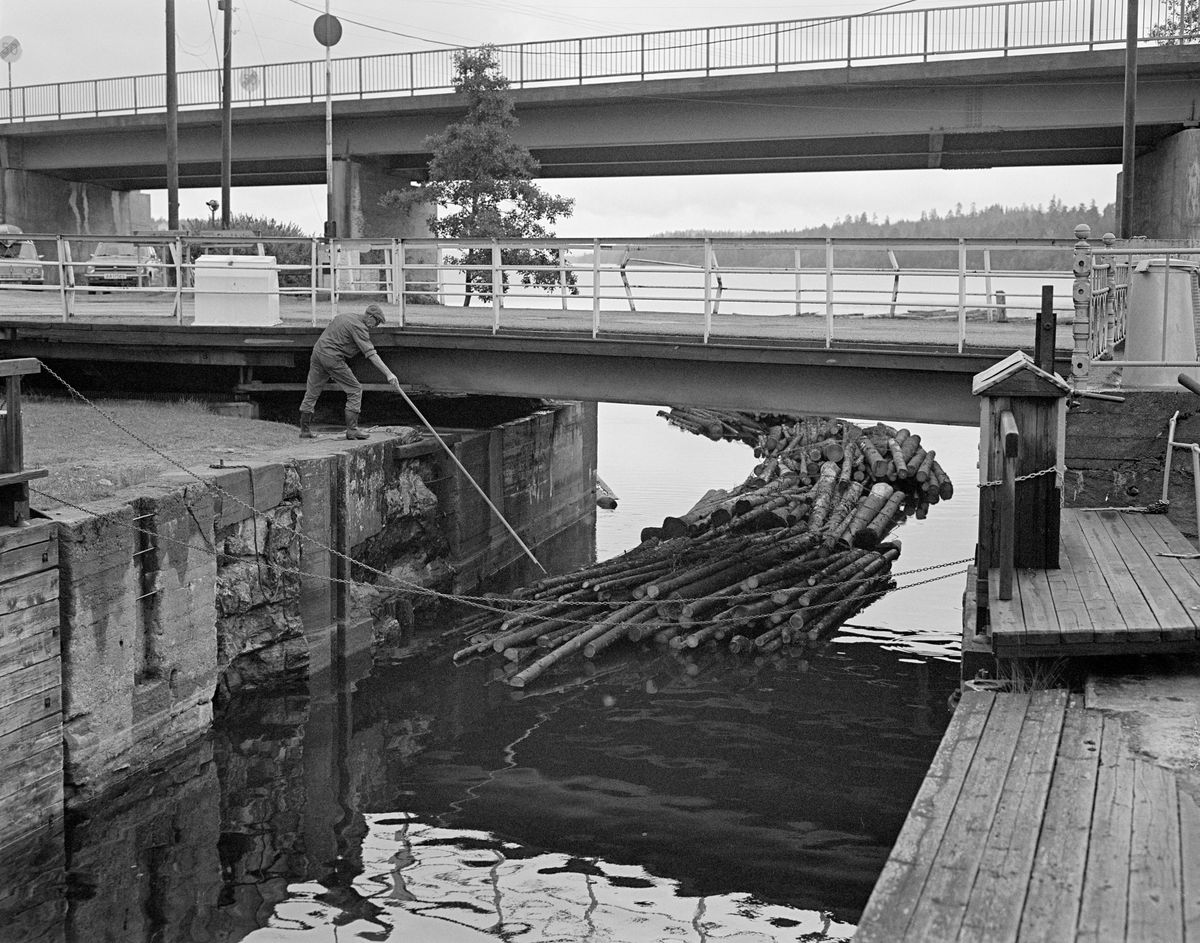 Fra sluseanlegget ved Ørje i Marker kommune i Østfold.  Fotografiet er tatt i 1982, som var den siste sesongen det ble fløtet tømmer på Haldenvassdraget.  Fotografiet er tatt ved innløpet til det øverste slusekammeret.  Til venstre i bildet ser vi Kåre Granli (1921-1991), som ved hjelp av en fløterhake skjøv på den fremste tømmerbunten, antakelig for å hindre at den skulle legge seg mot veggen i kanalen eller slusekammeret.  Over kanalløpet ser vi (nærmest) den lave, dreibare kanalbrua, bakenfor og høyere oppe E18-brua.  I perioden 1979-1982 ble det utelukkende fløtet ubarket massevirke i tre meters lengder i Haldenvassdraget.  Tømmeret ble utislått i bunter ved Skulerud i Høland.  Derfra ble det først fløtet i store slep til Ørje ved sørenden av Rødenessjøen.  Der måtte slepene deles opp i lenker som var tilpasset lengden på de tre slusekamrene.  Ved Ørje skulle tømmeret sluses 10 høydemeter ned, fra Rødenessjøen ovenfor til Ørjeelva nedenfor.  Med det nevnte virkessortimentet gikk det fire lastebilbunter i hver slusevending.

En liten historikk om tømmerfløting og kanaliseringsarbeid i Haldenvassdraget finnes under fanen «Opplysninger».