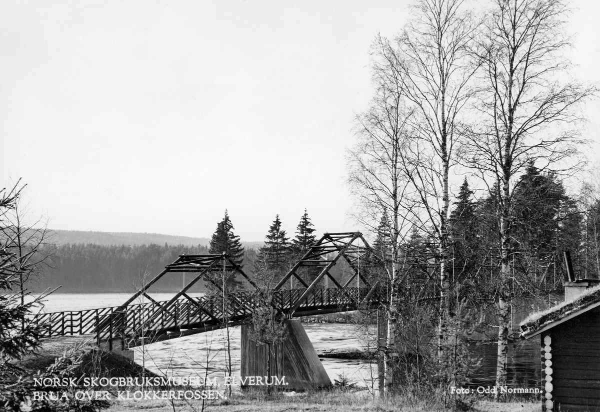Brua over Klokkerfossen i Elverum, mellom klokkergarden Fossum og Prestøya.  Fotografiet er tatt fra elvas østre bredd.  Brua ble bygd i 1964, ti år etter at nyetablerte Norsk Skogbruksmuseum fikk overta Prestøya som areal for sitt friluftsmuseum, og et drøyt år etter at museet fikk overta klokkergarden Fossum med 76 dekar jord og gjenstående bygninger.  Brua kvilte på støpte betongkar, ett på hver landside og fem ute i fossen.  Ute i elveløpet fikk den fire spenn a 20 meter.  Spennene mot land på begge sider var 14 meter lange.  Dermed ble brua 108 meter lang.  På de nevnte fundamentene ble det reist ei bru i tømmersprengverkskonstruksjon.  Brubygginga kostet 160 000 kroner i 1964.  Konservator Tore Fossum, som var museets eneste ansatte, var meget fornøyd med brua som skulle gjøre det lett å ferdes mellom nyetablerte kontorer og utstillinger på klokkergarden Fossum og friluftsmuseet på Prestøya: «Den har fått en estetisk meget god utforming og ligger godt i terrenget og blir godt likt av alle besøkende.  Den binder Prestøya sammen med Klokkergårdsarealet på en fin måte og forsterker samspillet mellom foss og skog, som er så viktige bakgrunnselementer for et skogbruks-, jakt- og fiskerimuseum.»  Helt til høyre i bildet er vi et hjørne av fløterkoia Fossheim.  Denne lå på Klokkergardens grunn og tilhørte Glomma fellesfløtningsforening, men ble overlatt til museet etter at fløtinga i vassdraget ble avviklet i 1985.