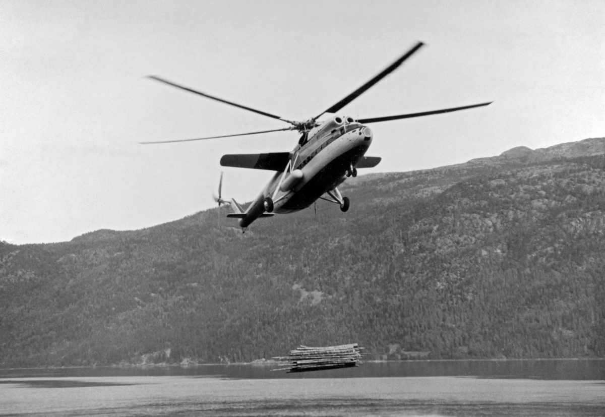Helikopter av typen MI-6, fotografert under nedstigning mot Kviteseidvatnet i Telemark under forsøk med tømmertransport fra det bratte fjellterrenget sør for Dyreskarnut ned til fløtingsvassdraget.  Den avbildete maskinen var i midten av 1960-åra verdens største helikopter.  Helikopterkroppen var 33,2 meter lang og rotorbladene spente over en diameter på 35 meter.  Maskinen veide 40,5 tonn og kunne i tillegg frakte 12 tonn nyttelast i et romslig, men enkelt utstyrt lasterom.  MI-6 hadde to kraftige gassturbinmotorer, som til sammen kunne utvikle 11 000 hestekrefter med et drivstofforbruk på 3 500 – 4 000 liter i timen.  I Kviteseid ble maskinen testet på henting av buntet tømmer fra den nevnte lokaliteten sør for Dyrskarnut, 4 860 meter fra og 390 meter høyere enn slippstedet i Kviteseidvatnet.  Tømmeret på hogstfeltet var på forhånd samlet i bunter med drøyt 5 tonns vekt og gjennomsnittlig 8,5 kubikkmeters volum (fastmasse).  Buntene ble hengt under helikopteret i cirka 10 meter lange bærestropper.  Konklusjonen fra disse undersøkelsene ble at kostnadsbildet minte om det man hadde hatt da man et par år tidligere hentet noe tømmer fra en annen lokalitet i området med et noe mindre helikopter et par år tidligere.  Den store russiske maskinen kunne riktignok ta noe større bunter, men på kløftet, blokkrik mark som her innebar det tidkrevende manuelt slit å samle store tømmerbunter.  «Selv om selve helikoptertransporten blir noe billigere med en større maskin, vil de øvrige kostnader bli noe høyere», skrev forsøkslederen, professor Ivar Samset (1918-2015).  Det han ikke skrev, men erfarte, var at helikoptertransport av tømmer var tapsbringende.  Driftskostnadene knyttet til arbeidet på hogstfeltet og bruken av helikopteret oversteg tømmerets omsetningsverdi.  Mer informasjon om forsøkene med helikoptertransport av tømmer fra vanskelig terreng finnes under fanen «Opplysninger».