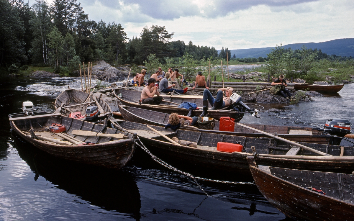 Kvilestund for fløterlaget som var ansvarlig for «sluttrensken» under fløtinga i Glomma gjennom Solør i 1985.  Fotografiet er tatt på et av steinskjærene i Norsfossen i Brandval, der elva, etter å ha flytt stille i over tre mil, passerte to terskler som ved lavvann skapte et fall på drøyt 3 meter.    Dette fotografiet viser hvordan tømmerfløterne, på en dag med lav vannføring, brukte denne lokaliteten som kvilested.  Vi ser til sammen sju tjærebredde robåter av tre med påhengsmotorer.  Et par av fløterne kvilte i båtene, men størstedelen av gruppa samlet seg på det nevnte steinskjæret, der vi også skimter en skådam av tre.