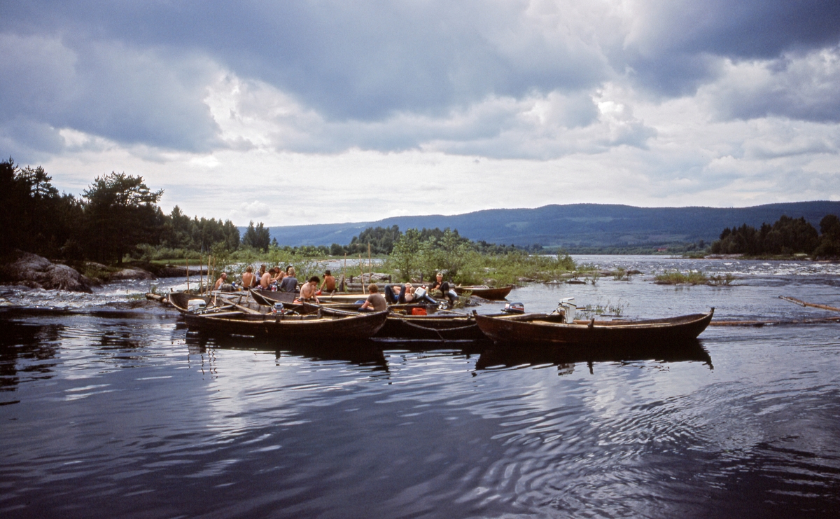 Kvilestund for fløterlaget som var ansvarlig for «sluttrensken» under fløtinga i Glomma gjennom Solør i 1985.  Fotografiet er tatt på et av steinskjærene i Norsfossen i Brandval, der elva, etter å ha flytt stille i over tre mil, passerte to terskler som ved lavvann skapte et fall på drøyt 3 meter.    Dette fotografiet viser hvordan tømmerfløterne, på en dag med lav vannføring, brukte denne lokaliteten som kvilested.  Vi ser til sammen sju tjærebredde robåter av tre med påhengsmotorer.  Et par av fløterne kvilte i båtene, men størstedelen av gruppa samlet seg på det nevnte steinskjæret, der vi også skimter en skådam av tre.