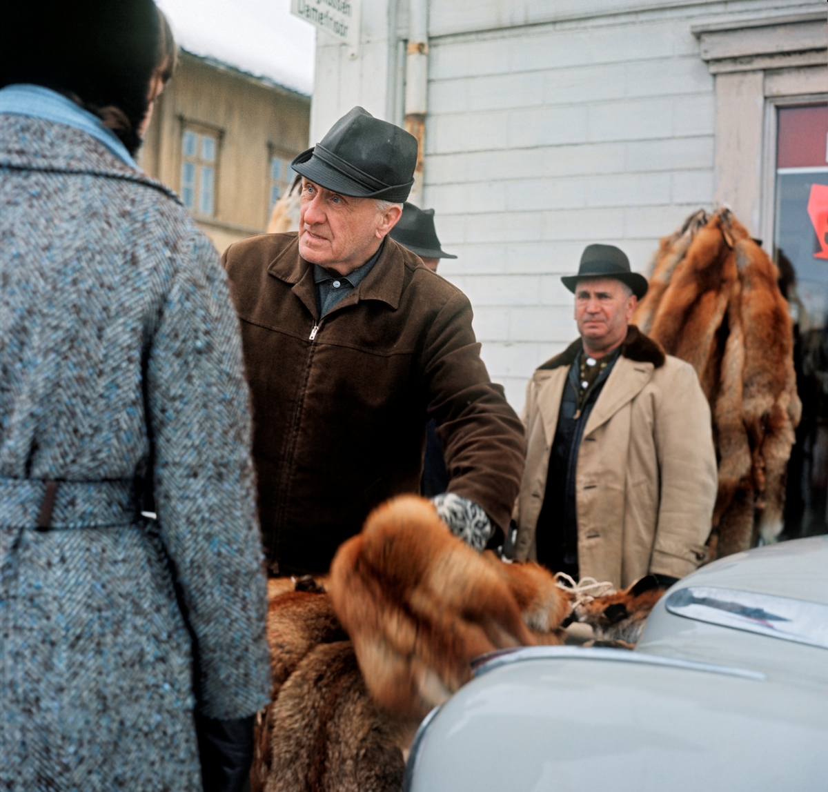 Skinnhandel under Grundsetmart’n i Leiret, kommunesenteret i Elverum, i mars 1970.  Fotografiet er tatt utenfor den bygningen som har vært kalt "Bekkevoll", "Bekkevold" eller "Bækkevold", der øst-vestgående og nord-sørgående gjennomfartsveg møttes.  Gustav Rogstad (1904-1978) drev til vanlig en liten klesforretning, men under mart’n trakk han ut på fortauet der han opptrådte som skinnoppkjøper.  På dette bildet ser vi ham (til høyre) mens jergeren Leiv Glad Ørbak (1897-1990) viste fram skinn på et bord som sto framfor en parkert bil.  Stundom brukte Rogstad også bilpanseret som disk under besiktigelsen av skinn han ble tilbudt.  Bilen skulle nok også få interesserte til å stanse slik at Rogstad fikk kontakt med dem.  Da dette bildet ble tatt var det ei kvinne (til venstre, med ryggen mot fotografen) som hadde stanset og fikk litt informasjon om skinnvarene.