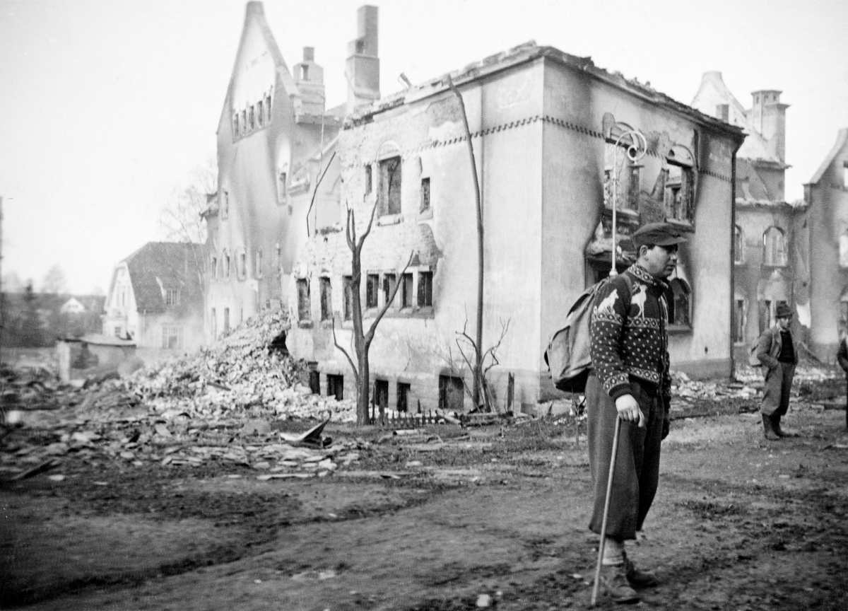 Lokalbefolkningen i Elverum inspiserer ruinene etter den tyske bombinga av kommunesenteret Leiret 11. april 1940.  At Elverum ble rammet av krigen på denne måten skyldtes at Stortinget, regjeringen, kronprinsen og kongen flyktet nordover da den tyske invasjonen av hovedstaden Oslo skjedde om morgenen 9. april.  Etter et møte i Festiviteten på Hamar flykten de nevnte myndighetsrepresentantene videre til Elverum folkehøgskole seinere på dagen den 9. april.    Tyske militære forsøkte ved en dristig manøver å innhente dem, men ble stanset etter en trefning ved Midtskogen, noen få kilometer vest for tilfluktsstedet.  Den 10. april nådde den tyske sendemannen Curt Bräuer Elverum med sitt krav til konge og regjering om at Norge skulle kapitulere, og at Vidkunn Quisling skulle utnevnes til ny statsminister.  Kongen var umiddelbart negativ, og regjeringen avviste også etter hvert det tyske kravet.  Okkupasjonsmaktens reaksjon på dette ble bombing av Elverum sentrum, noe som la mye av bebyggelsen i ruiner.  Dette fotografiet er tatt i det som hadde vært St. Olavsgate, der to menn med ryggsekker betraktet de omfattende skadene.  Murveggene og pipene på kommunehuset Elvarheim sto, til tross for de påkjenningene trykket fra bombing og påfølgende brann hadde utløst. 

En beskrivelse av den dramatiskem bombinga av Leiret i 1940 finnes under fanen «Opplysninger».